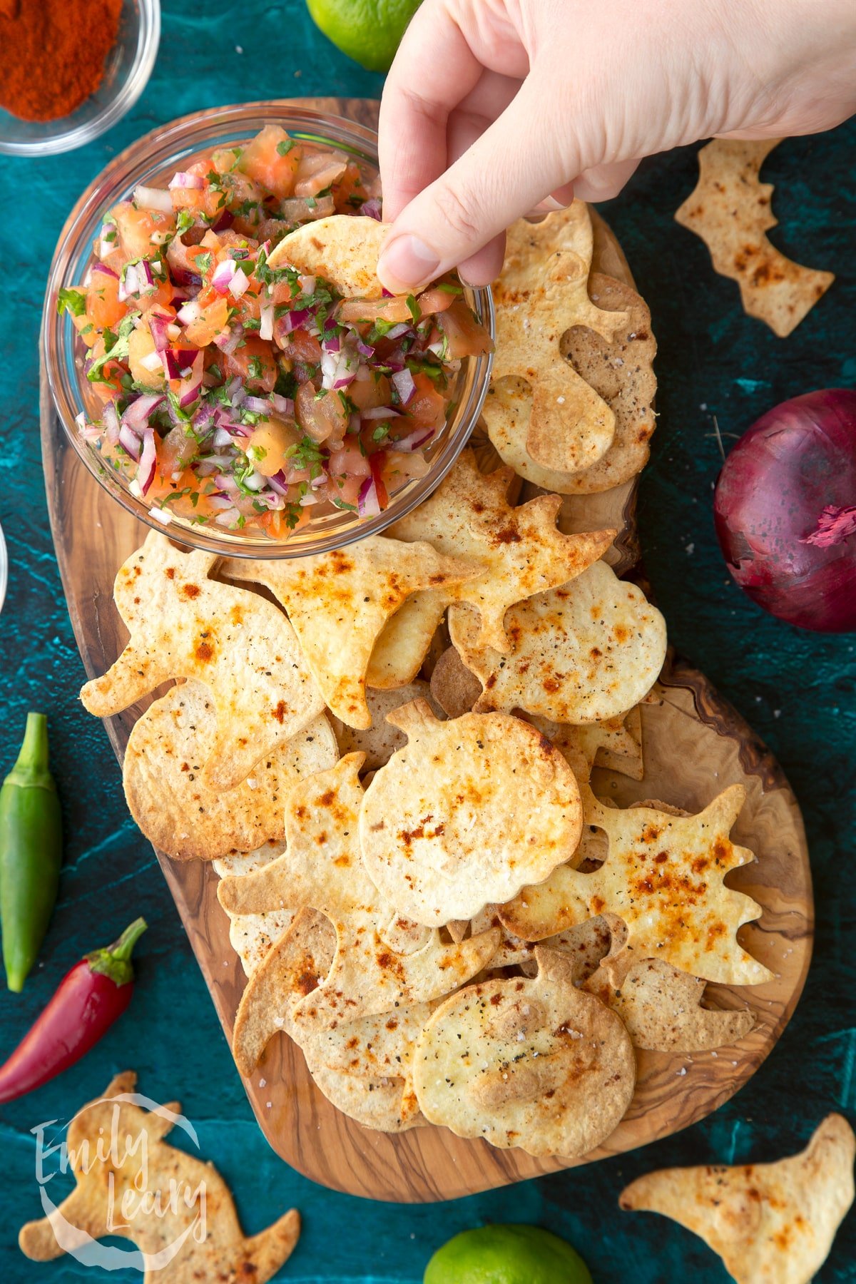 Halloween tortillas on a board with a small bowl of tomato salsa. A hand dips half a pumpkin shaped chip.