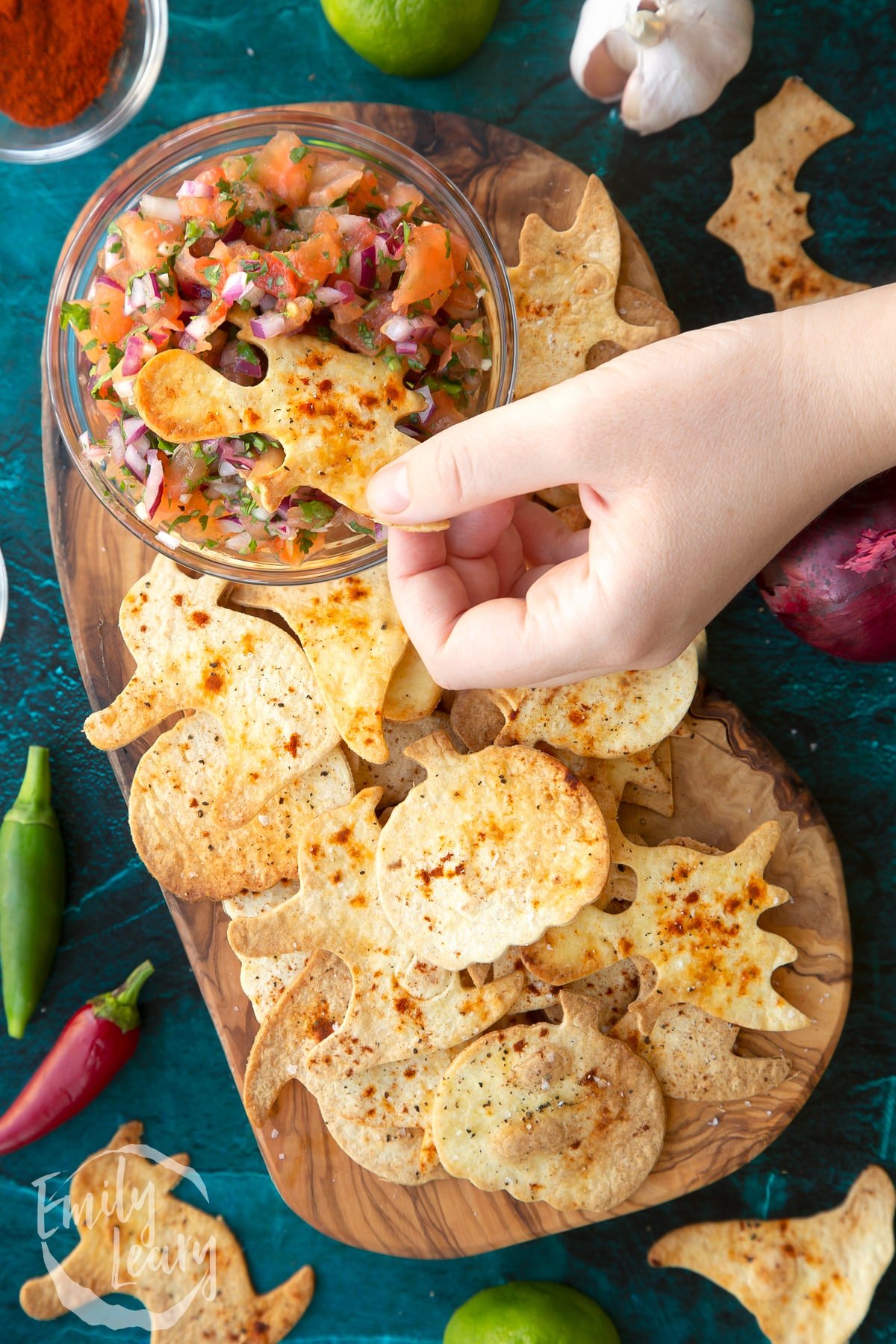 Halloween tortillas on a board with a small bowl of tomato salsa. A hand dips a ghost shaped chip.