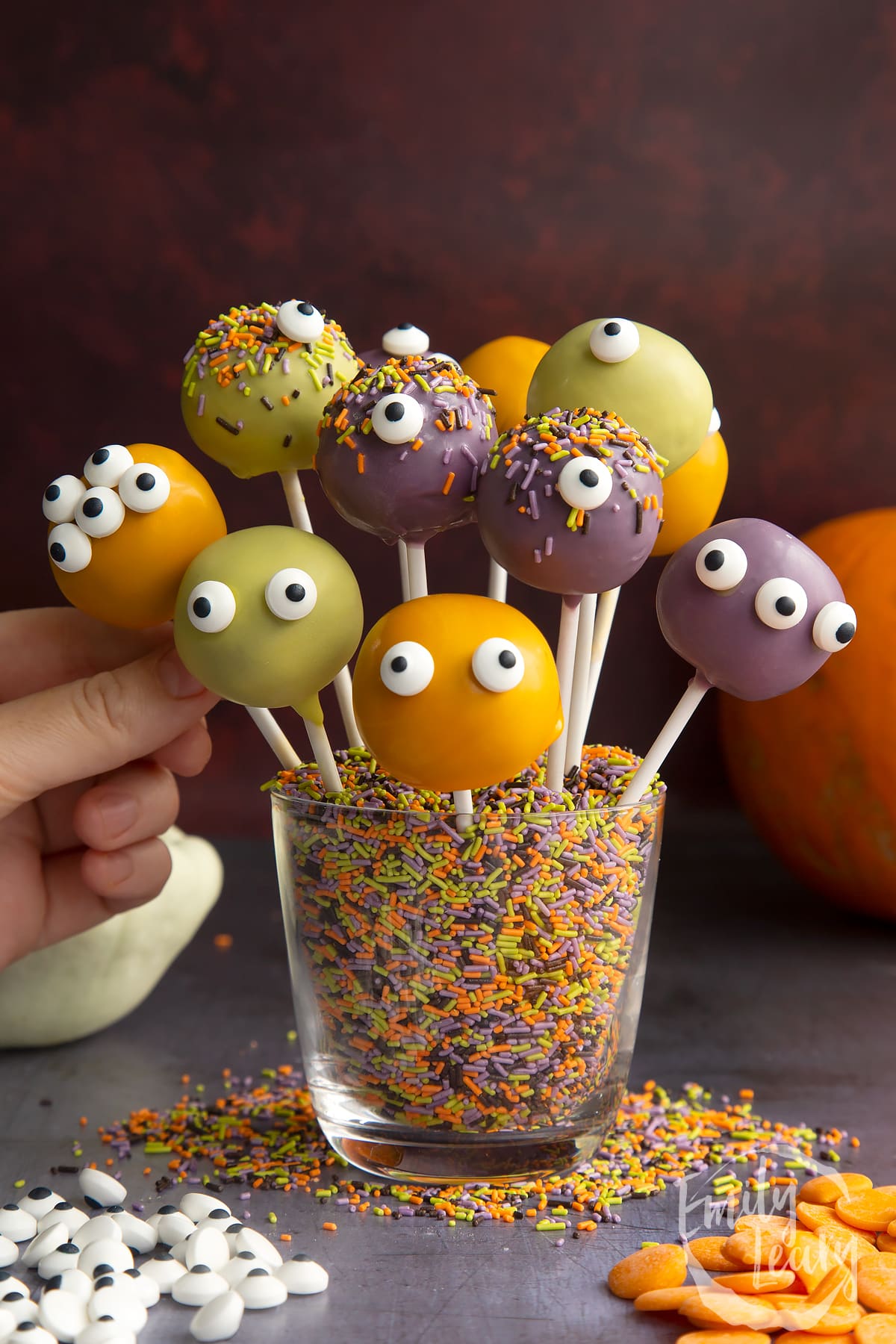 Halloween cake pops standing in a glass filled with Halloween sprinkles. A hand reaches for one.