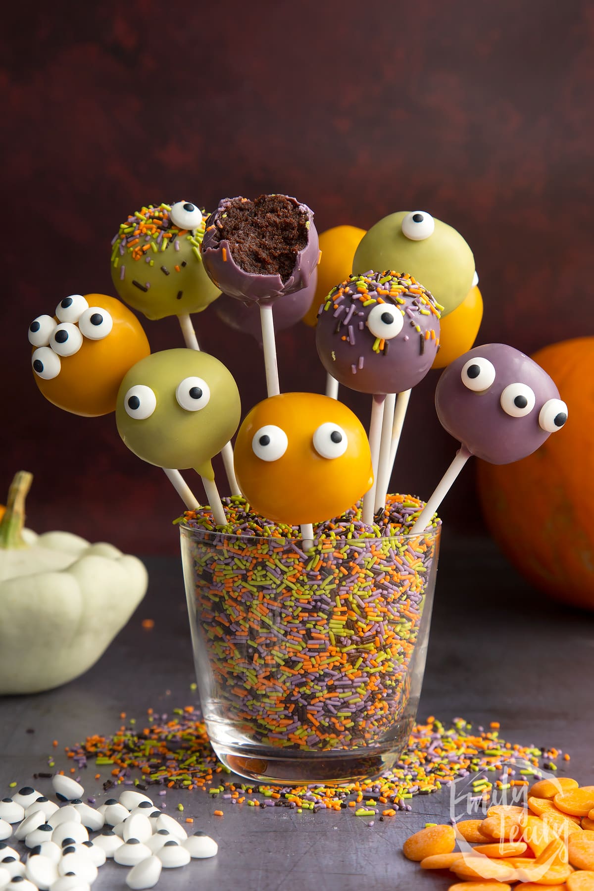 Halloween cake pops standing in a glass filled with Halloween sprinkles. One has been bitten open.