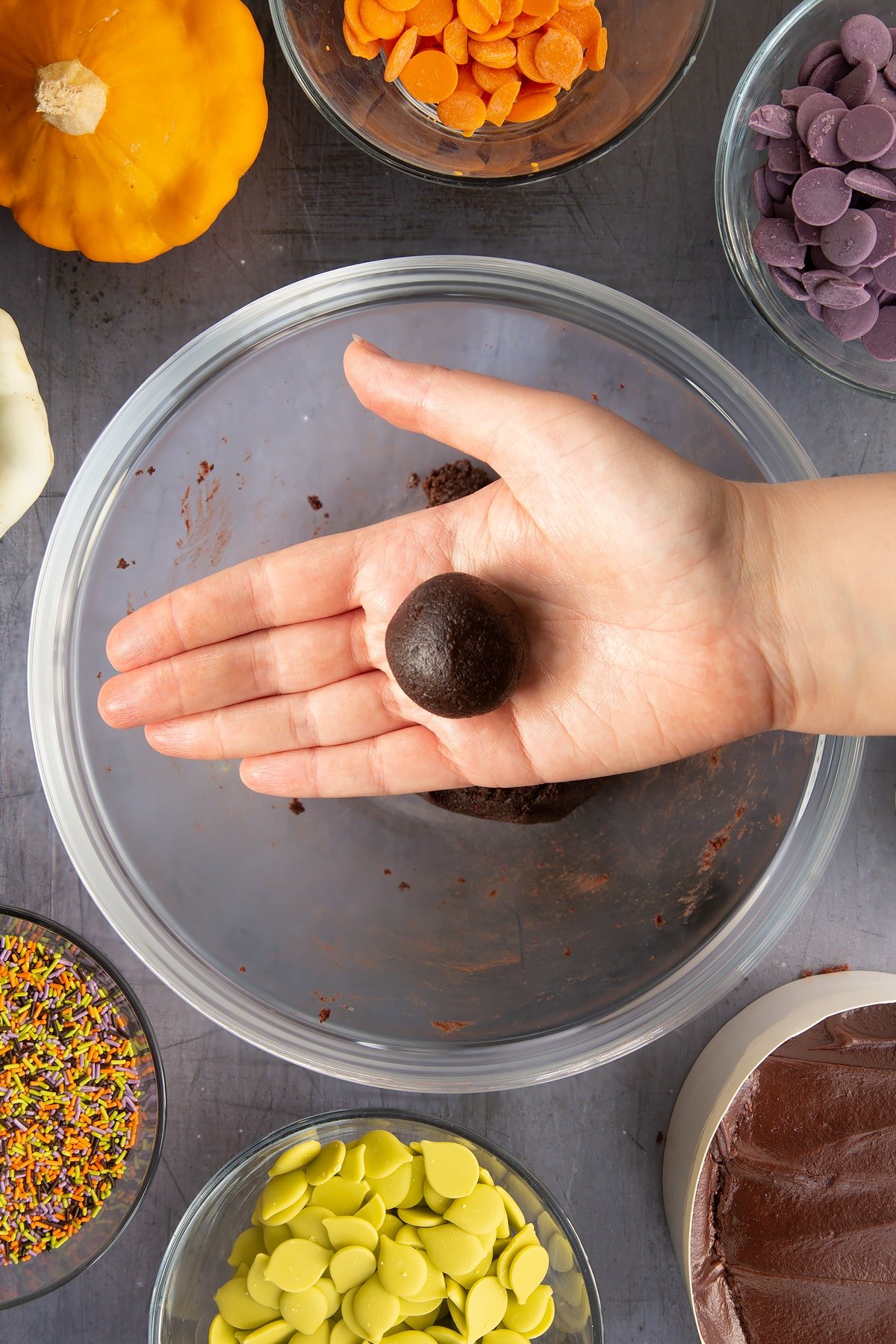 A hand holding a ball of chocolate cake. Ingredients to make Halloween cake pops surround the bowl beneath.