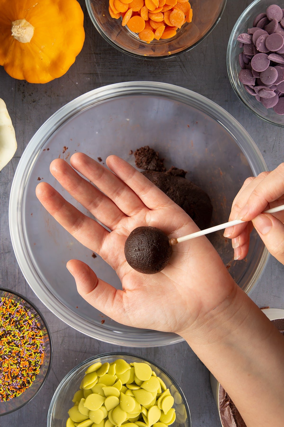 A hand holding a ball of chocolate cake. The other hand holds a stick with melted chocolate on the tip. Ingredients to make Halloween cake pops surround the bowl beneath.
