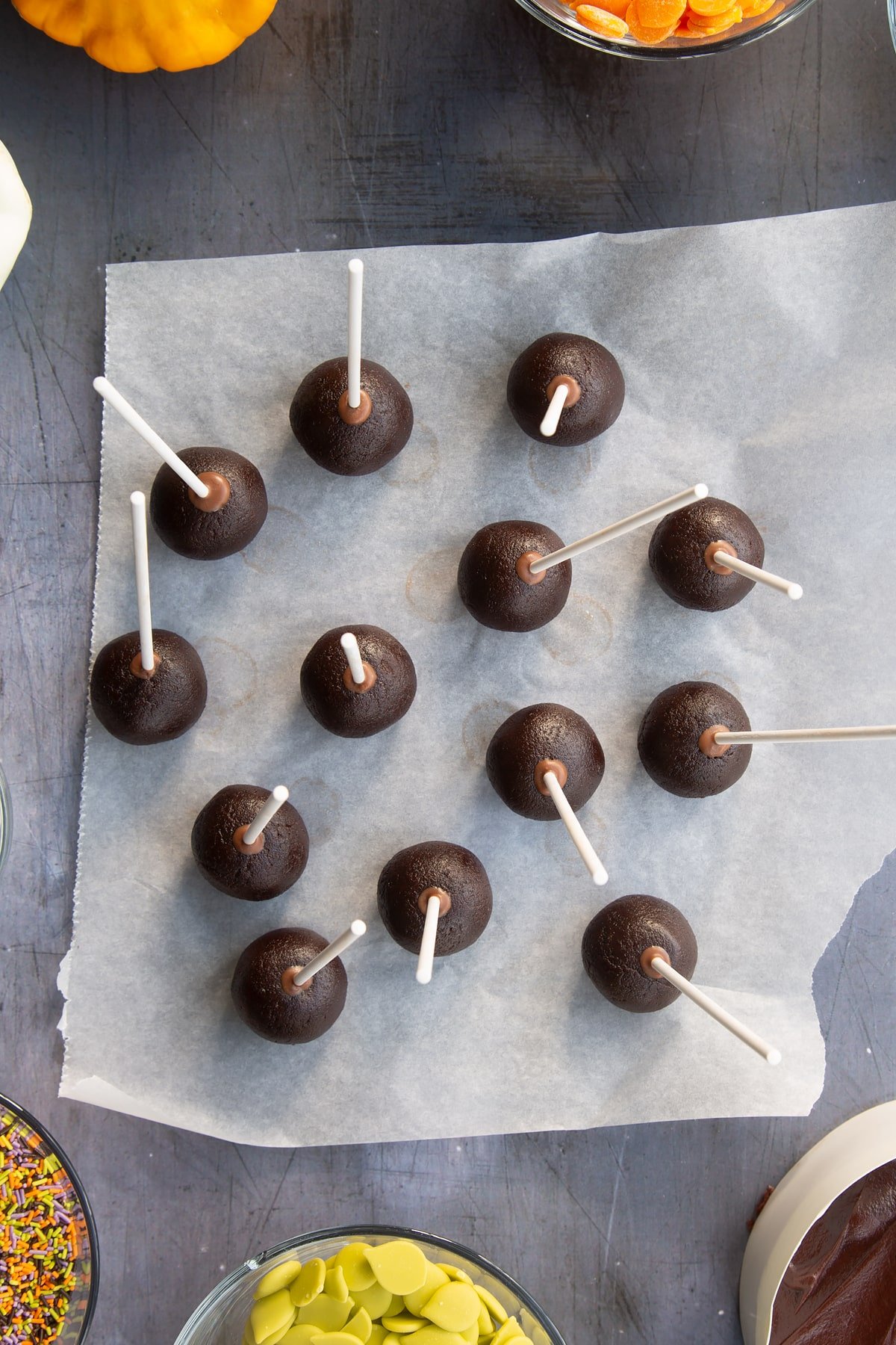 Uncovered cake pops with sticks, standing on a piece of baking paper. Ingredients to make Halloween cake pops surround the pan.