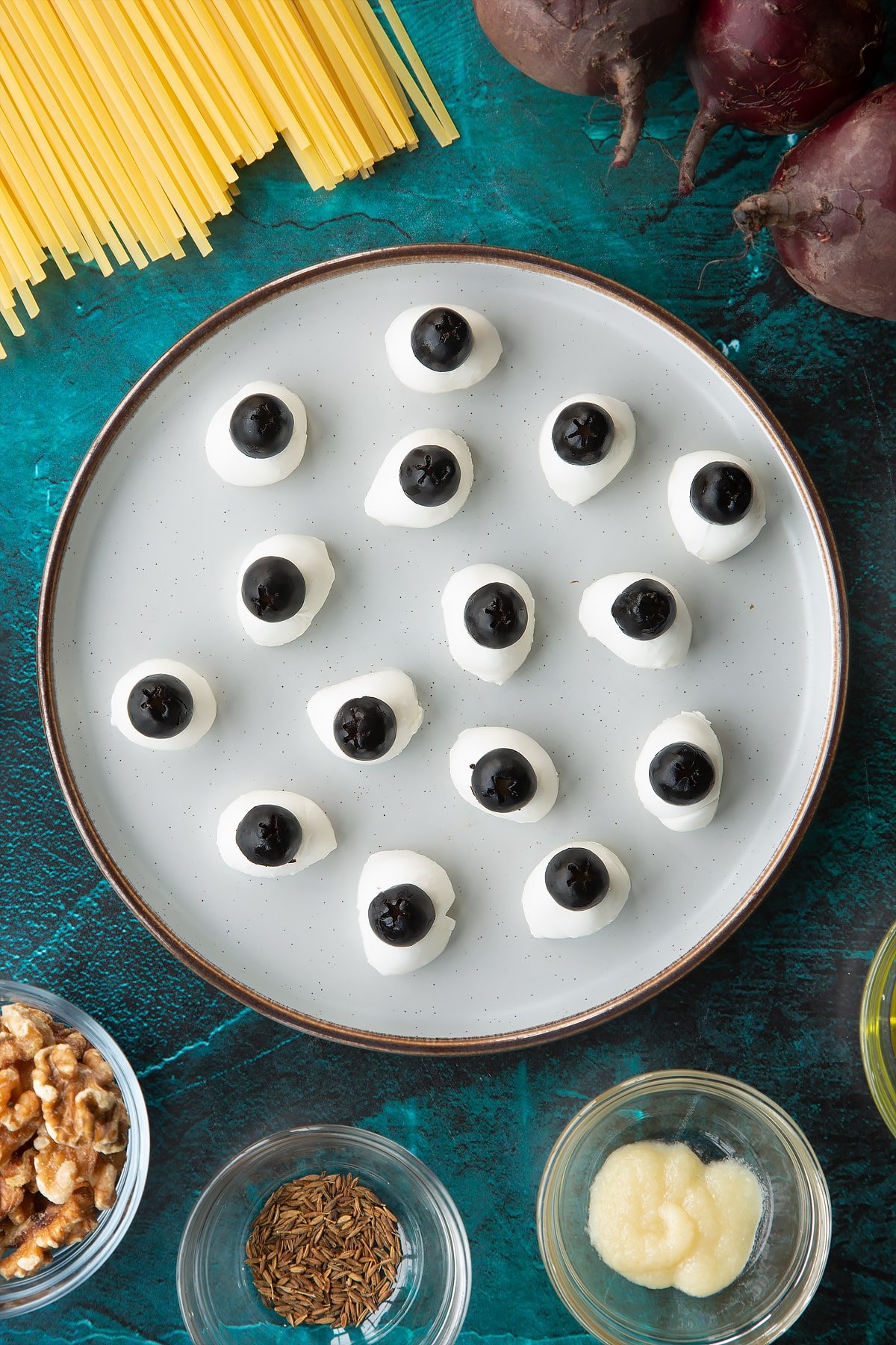 Mozzarella pearls topped with black olives on a plate. Ingredients to make a Halloween pasta recipe surround the plate.