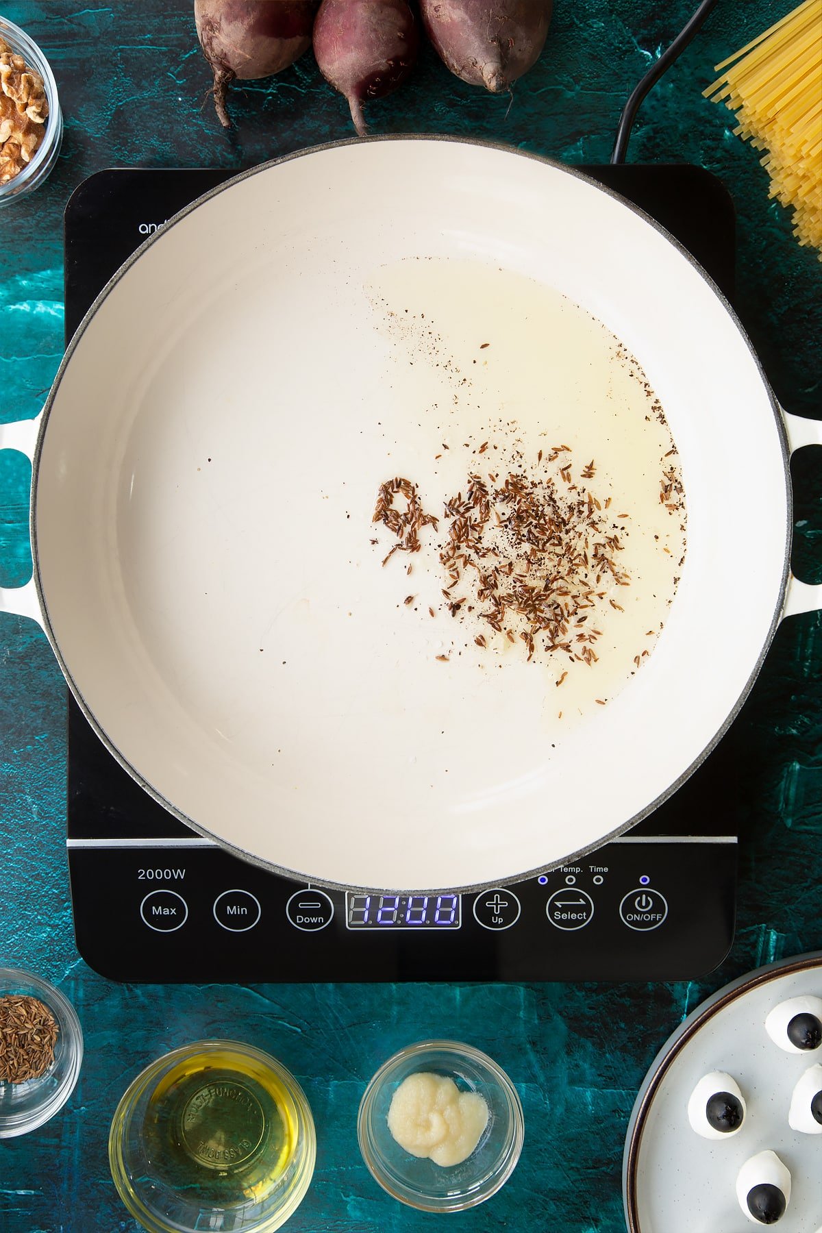 Fried cumin seeds in oil in a pan. Ingredients to make a Halloween pasta recipe surround the pan.