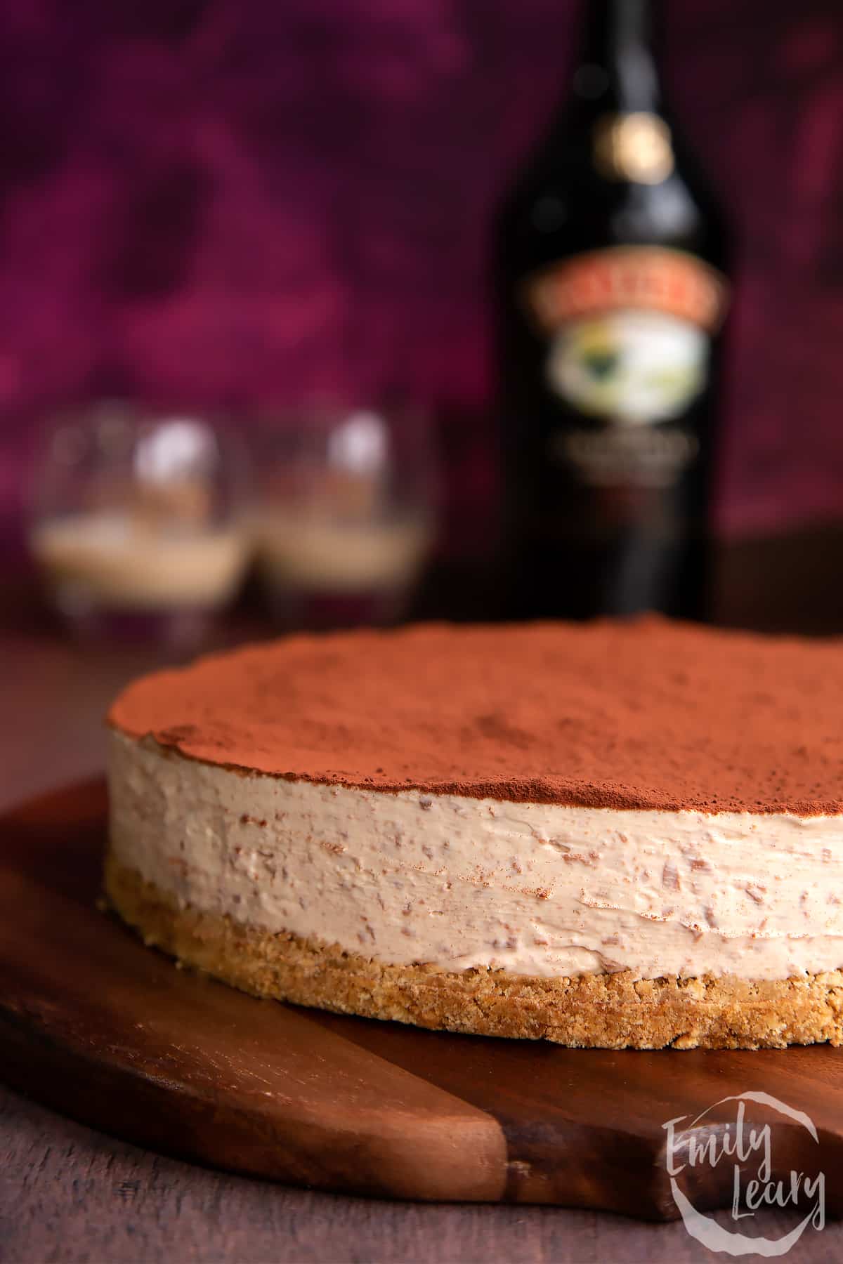 Baileys cheesecake topped with cocoa on a wooden board. A bottle of Baileys is in the background with two glasses.