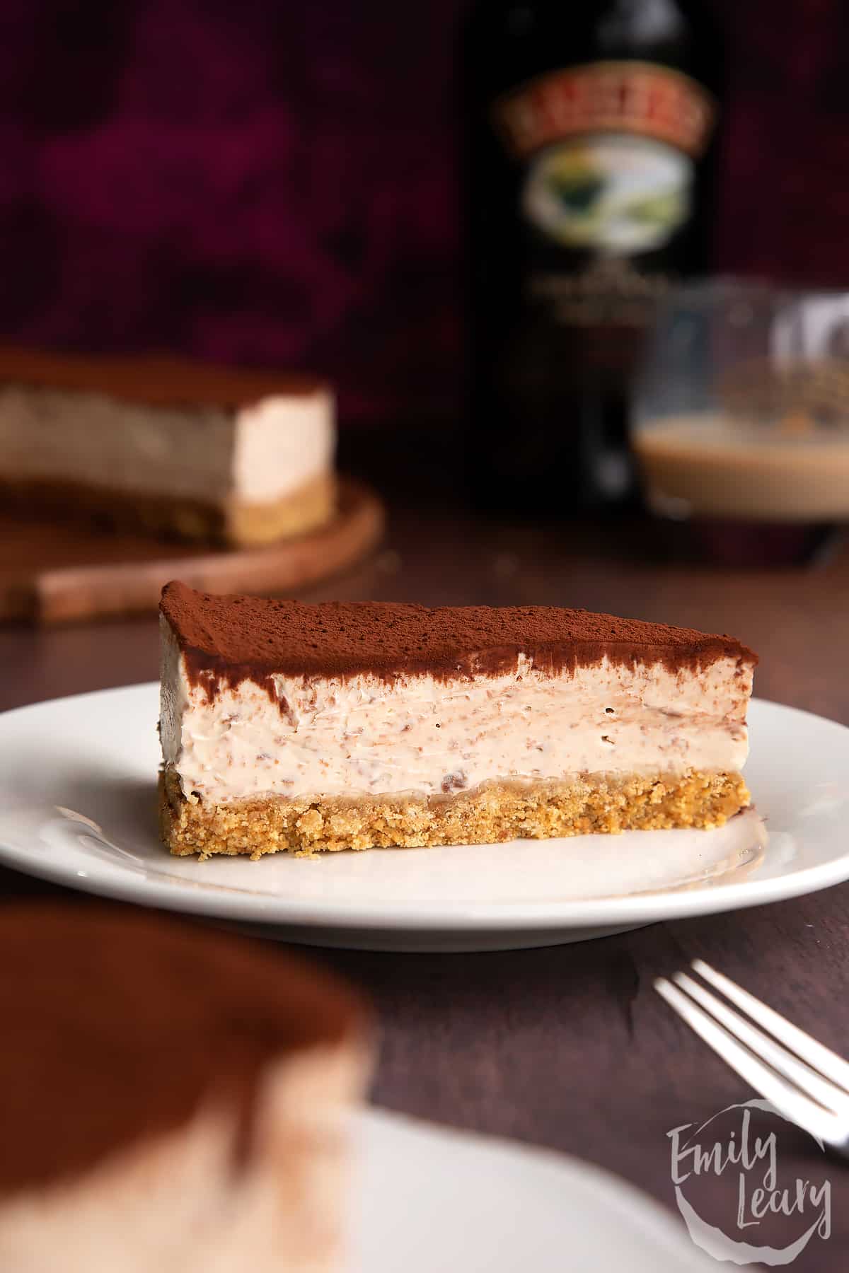 Slice of Baileys cheesecake topped with cocoa on a white plate. More cheesecake and a bottle of Baileys is visible in the background.