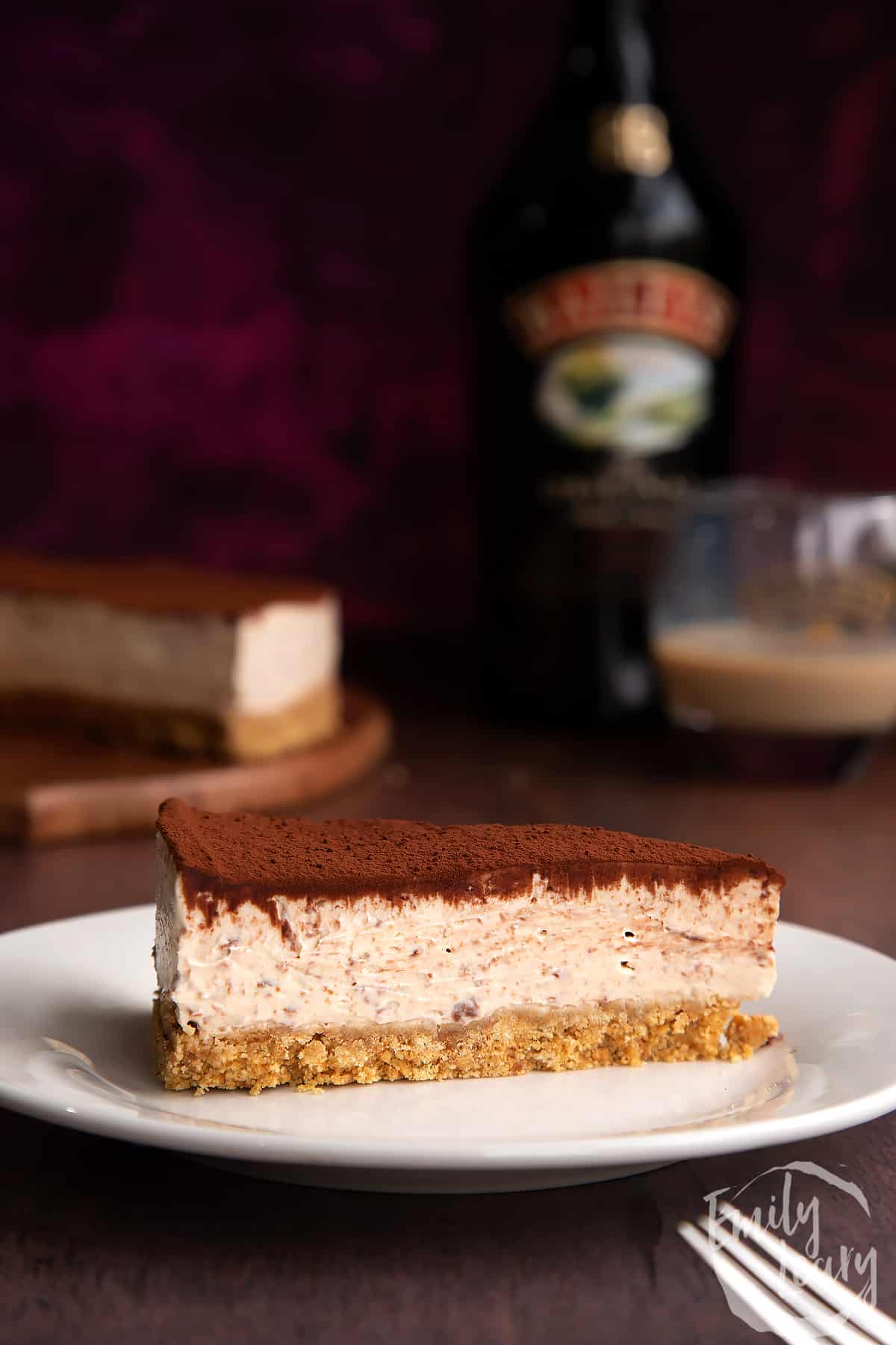 Slice of Baileys cheesecake topped with cocoa on a white plate. More cheesecake and a bottle of Baileys is in the background.