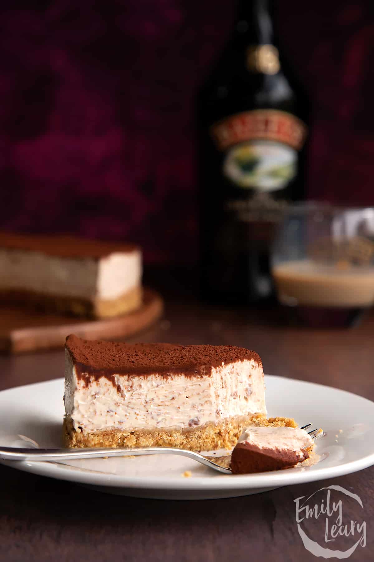 Slice of Baileys cheesecake topped with cocoa on a white plate. A fork rests with some cheesecake on it. More cheesecake and a bottle of Baileys is in the background.