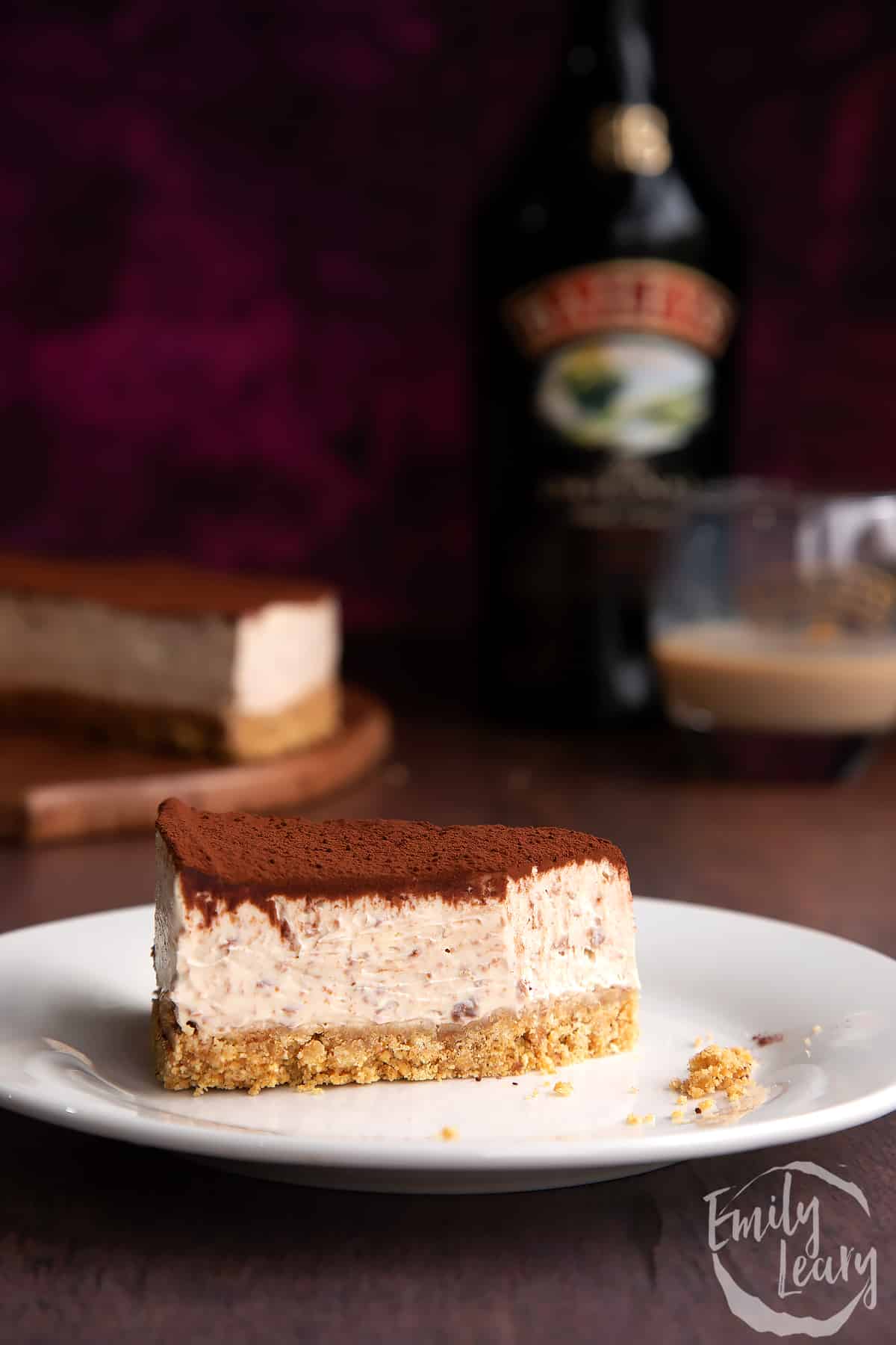 Slice of Baileys cheesecake topped with cocoa on a white plate. Part has been eaten. More cheesecake and a bottle of Baileys is in the background.