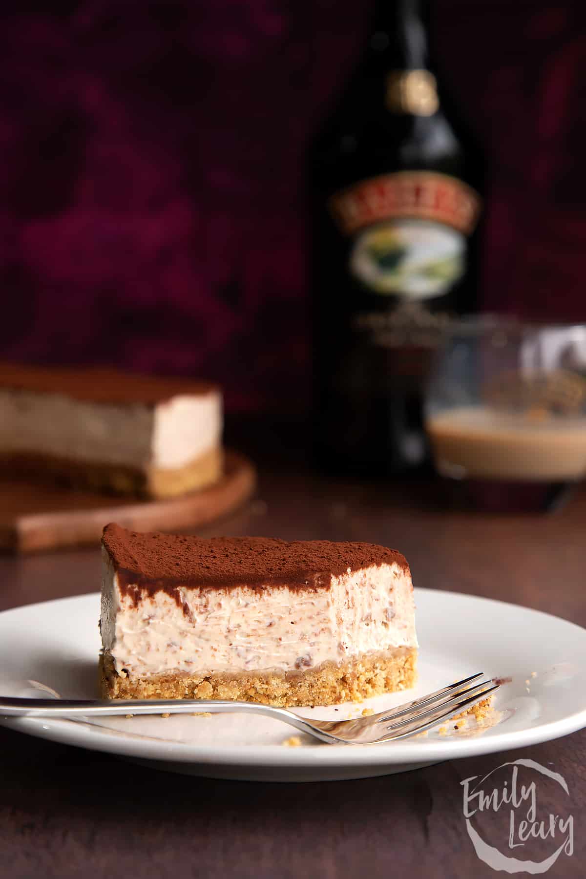 Slice of Baileys cheesecake topped with cocoa on a white plate. A fork rests in some crumbs. More cheesecake and a bottle of Baileys is in the background.