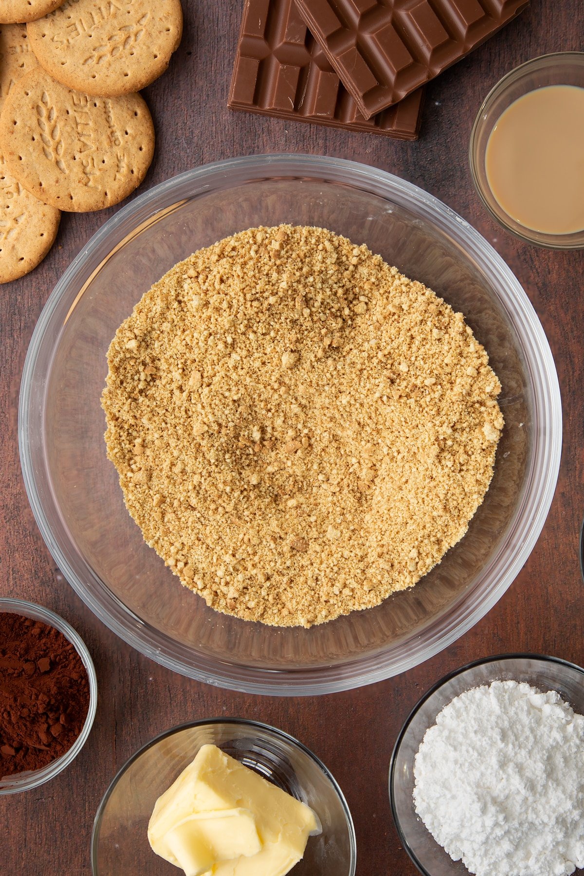 Digestive biscuit crumbs in a glass bowl. Ingredients to make Baileys cheesecake surround the bowl.