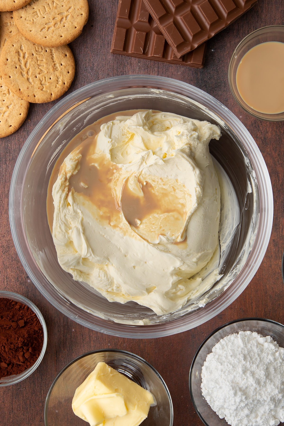 Whipped double cream, cream cheese and icing sugar in a glass bowl with Baileys on top. Ingredients to make Baileys cheesecake surround the bowl.