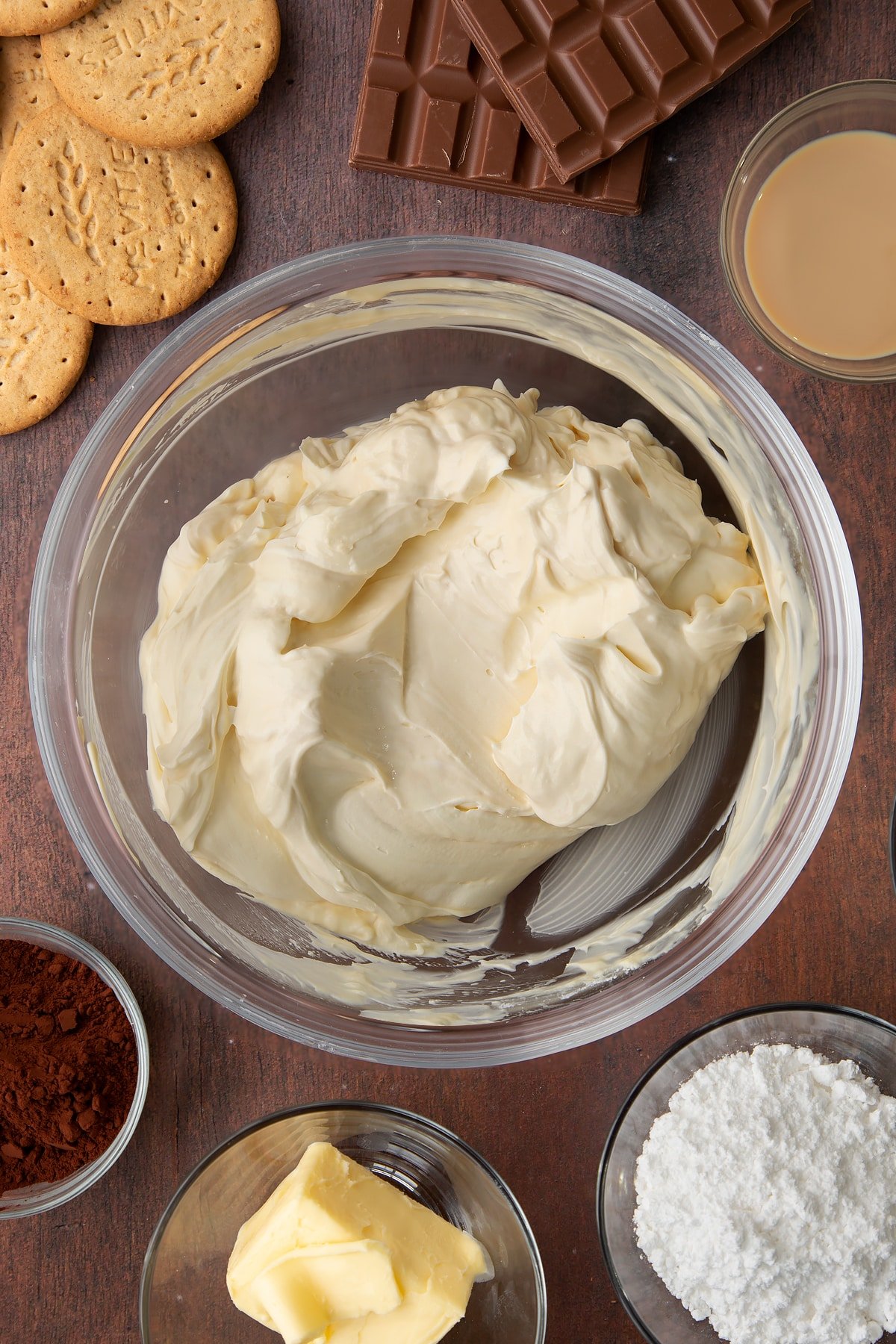 Whipped double cream, cream cheese, icing sugar and Baileys folded together in a glass bowl. Ingredients to make Baileys cheesecake surround the bowl.