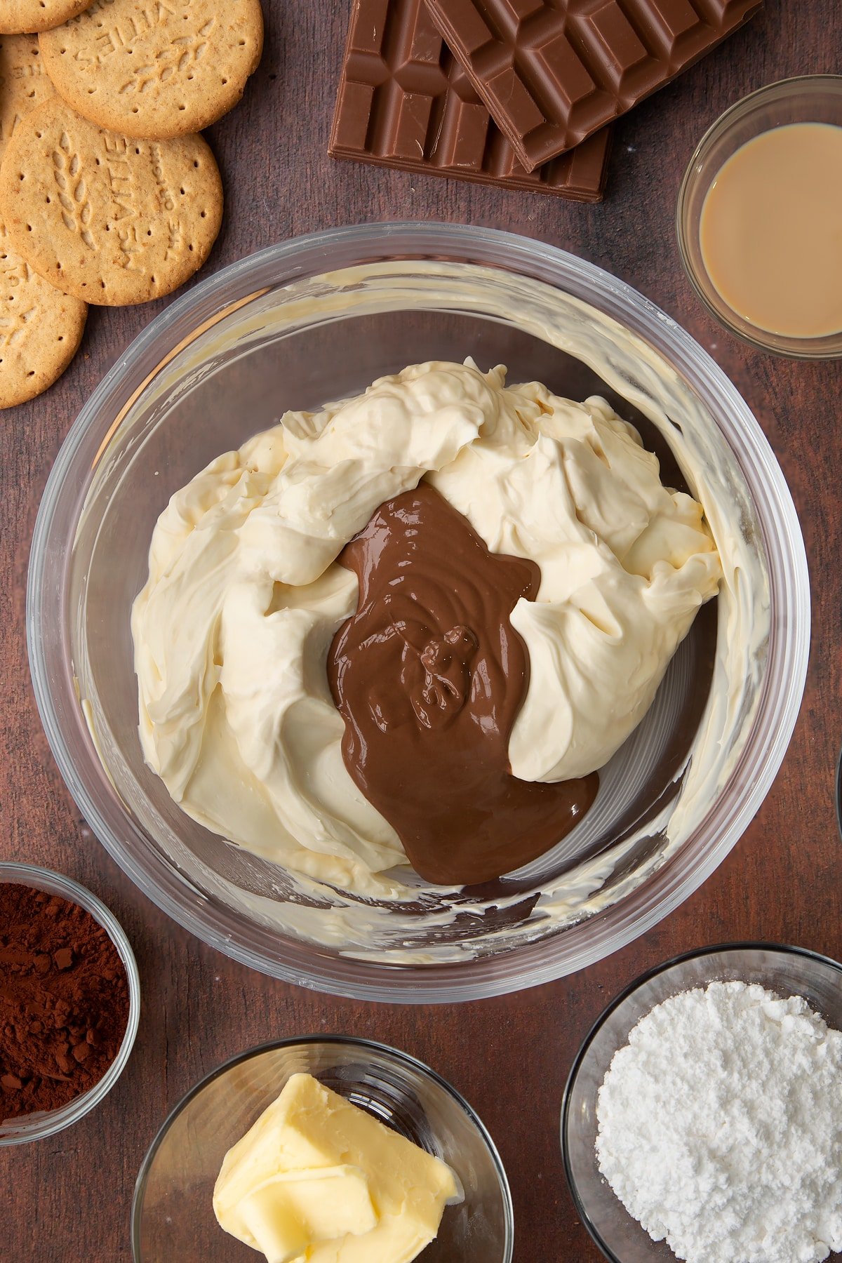 Whipped double cream, cream cheese, icing sugar and Baileys folded together with melted chocolate on top in a glass bowl. Ingredients to make Baileys cheesecake surround the bowl.