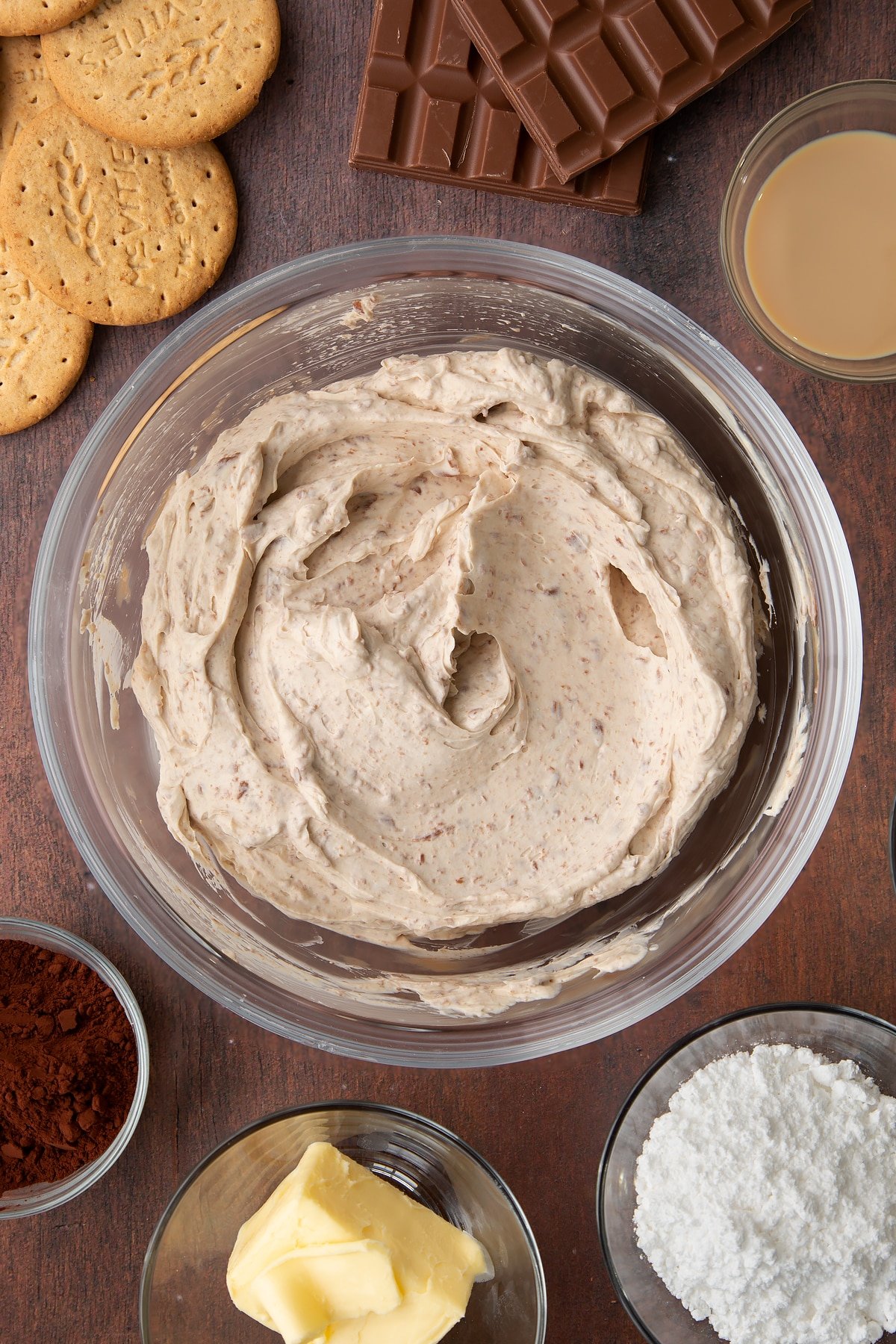 Whipped double cream, cream cheese, icing sugar, Baileys and melted chocolate folded together in a glass bowl. Ingredients to make Baileys cheesecake surround the bowl.