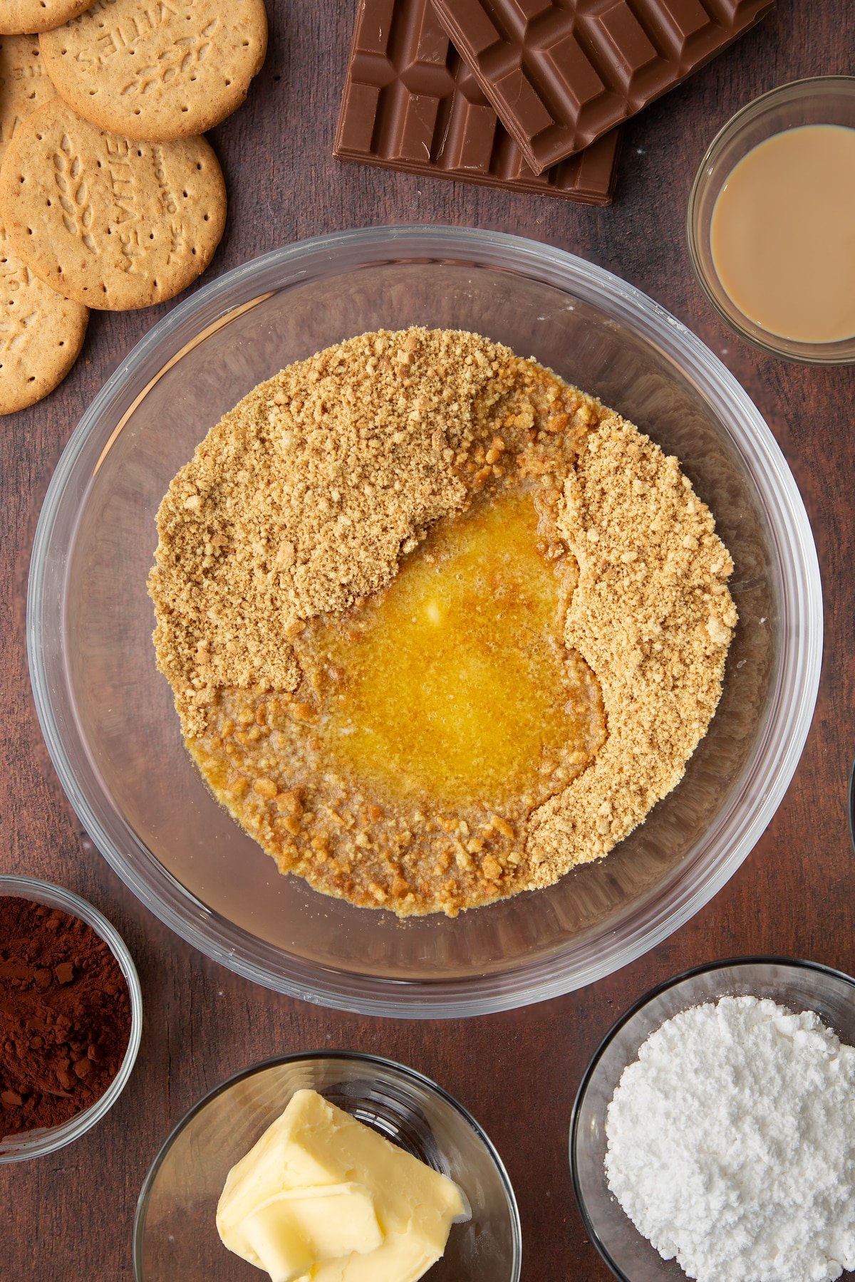 Digestive biscuit crumbs and melted butter in a glass bowl. Ingredients to make Baileys cheesecake surround the bowl.