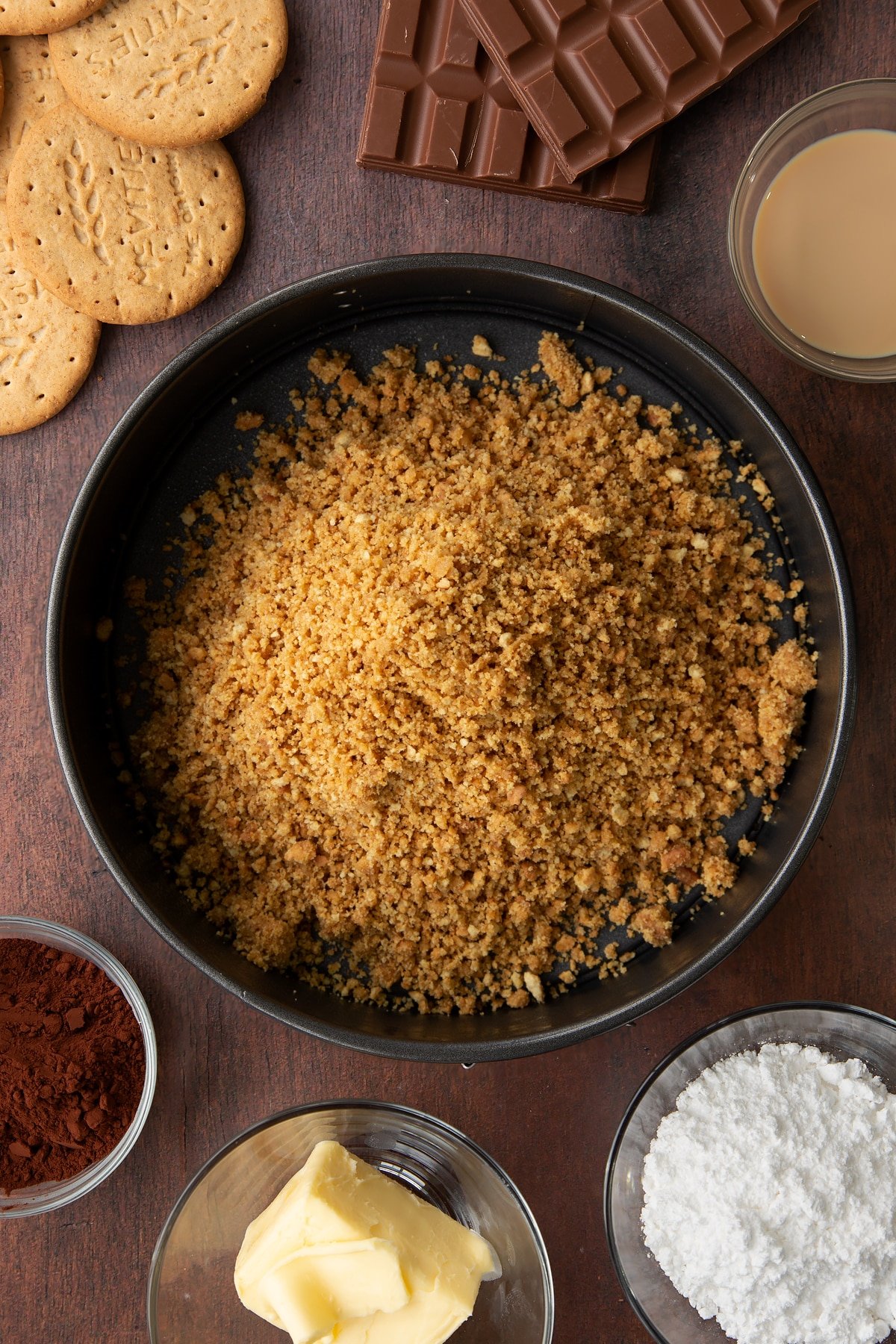 Digestive biscuit crumbs and melted butter mixed together in a cake tin. Ingredients to make Baileys cheesecake surround the tin.