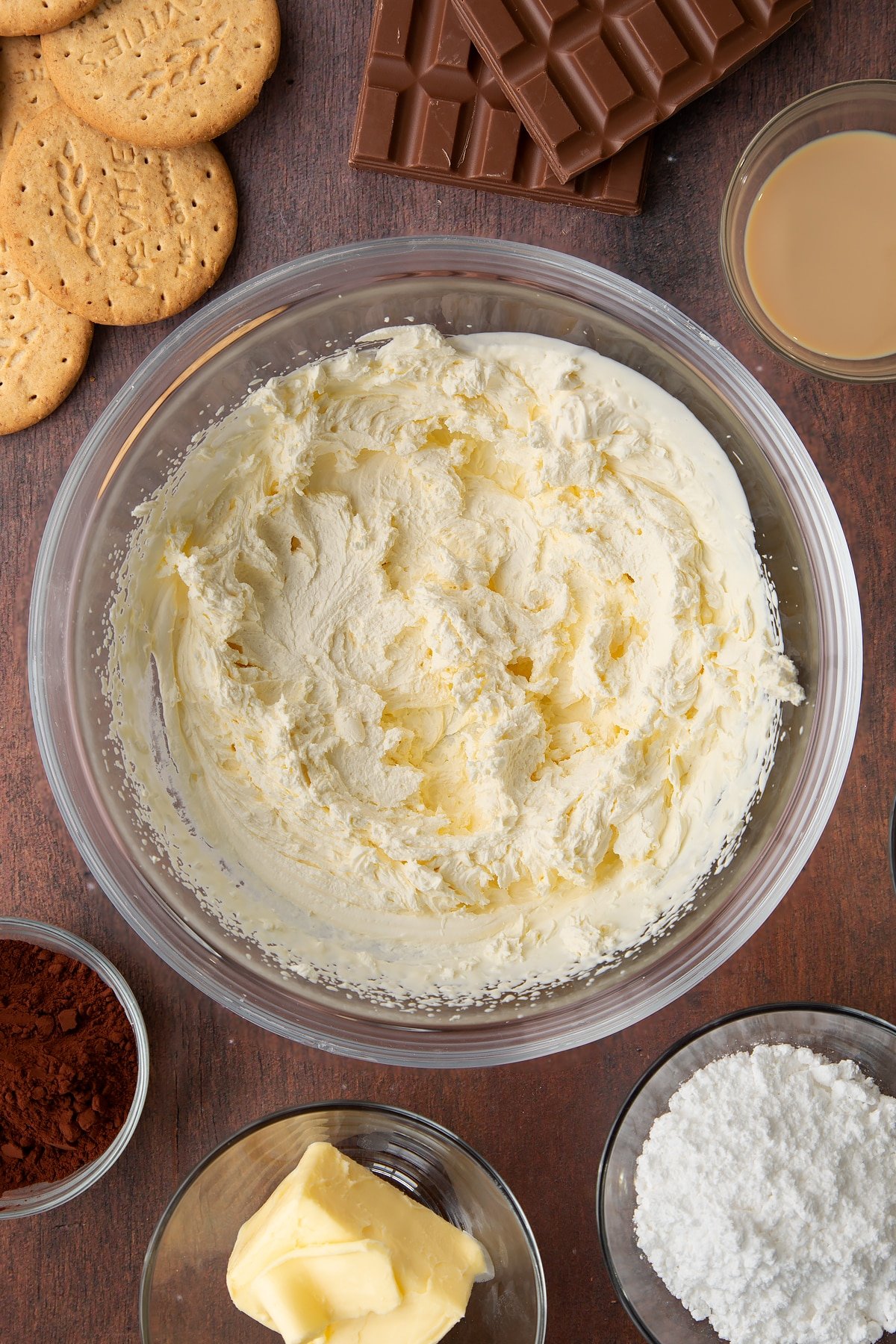 Whipped double cream in a glass bowl. Ingredients to make Baileys cheesecake surround the bowl.