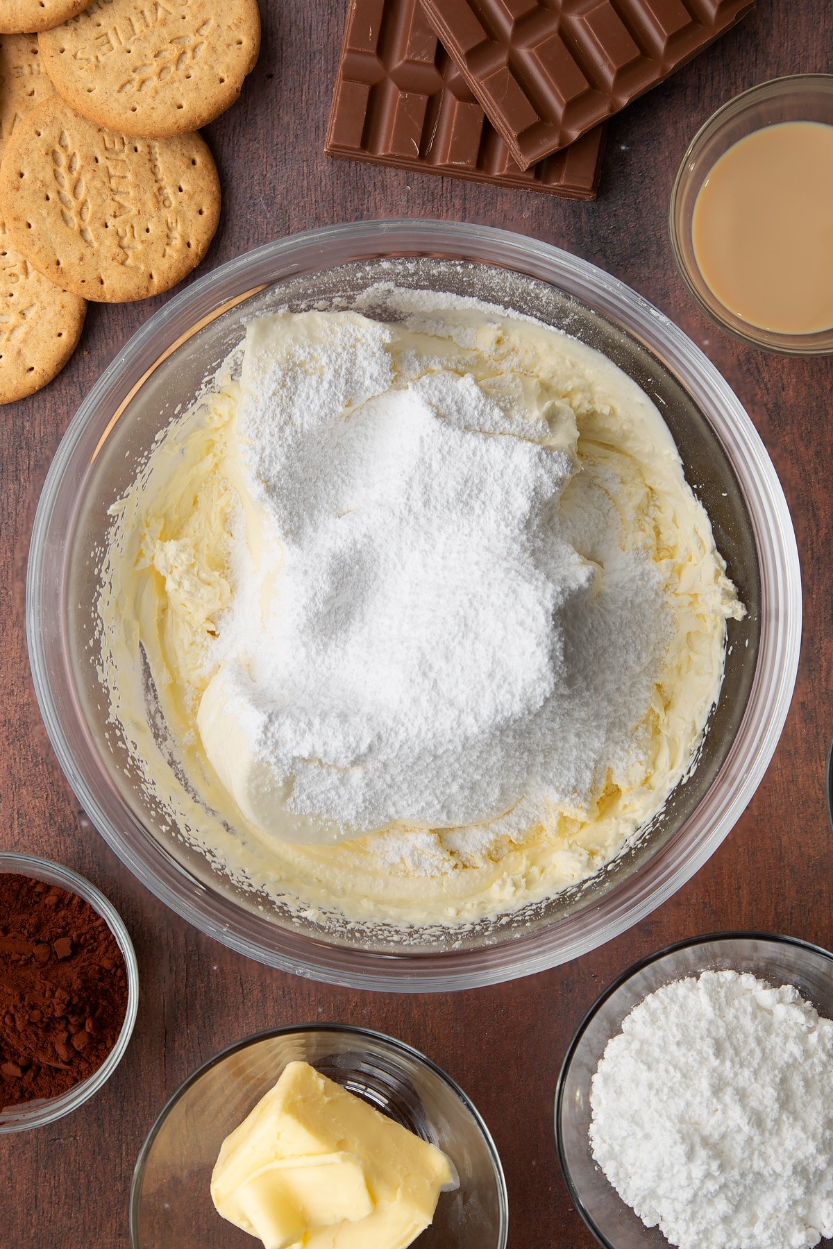 Whipped double cream, cream cheese and icing sugar in a glass bowl. Ingredients to make Baileys cheesecake surround the bowl.