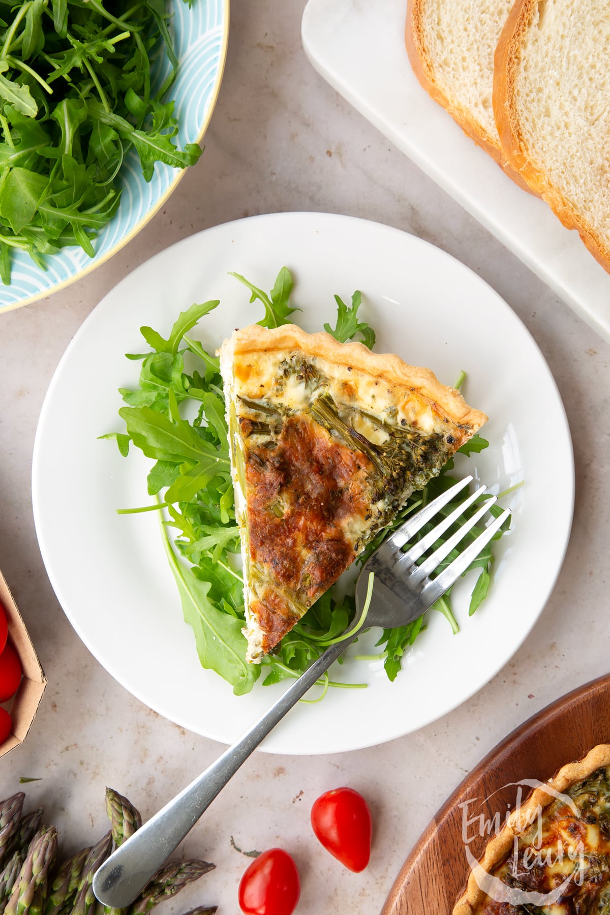 Overhead shot of the finished slice of broccoli and asparagus quiche served on a white plate ontop of a bed of salad with a fork on the side.