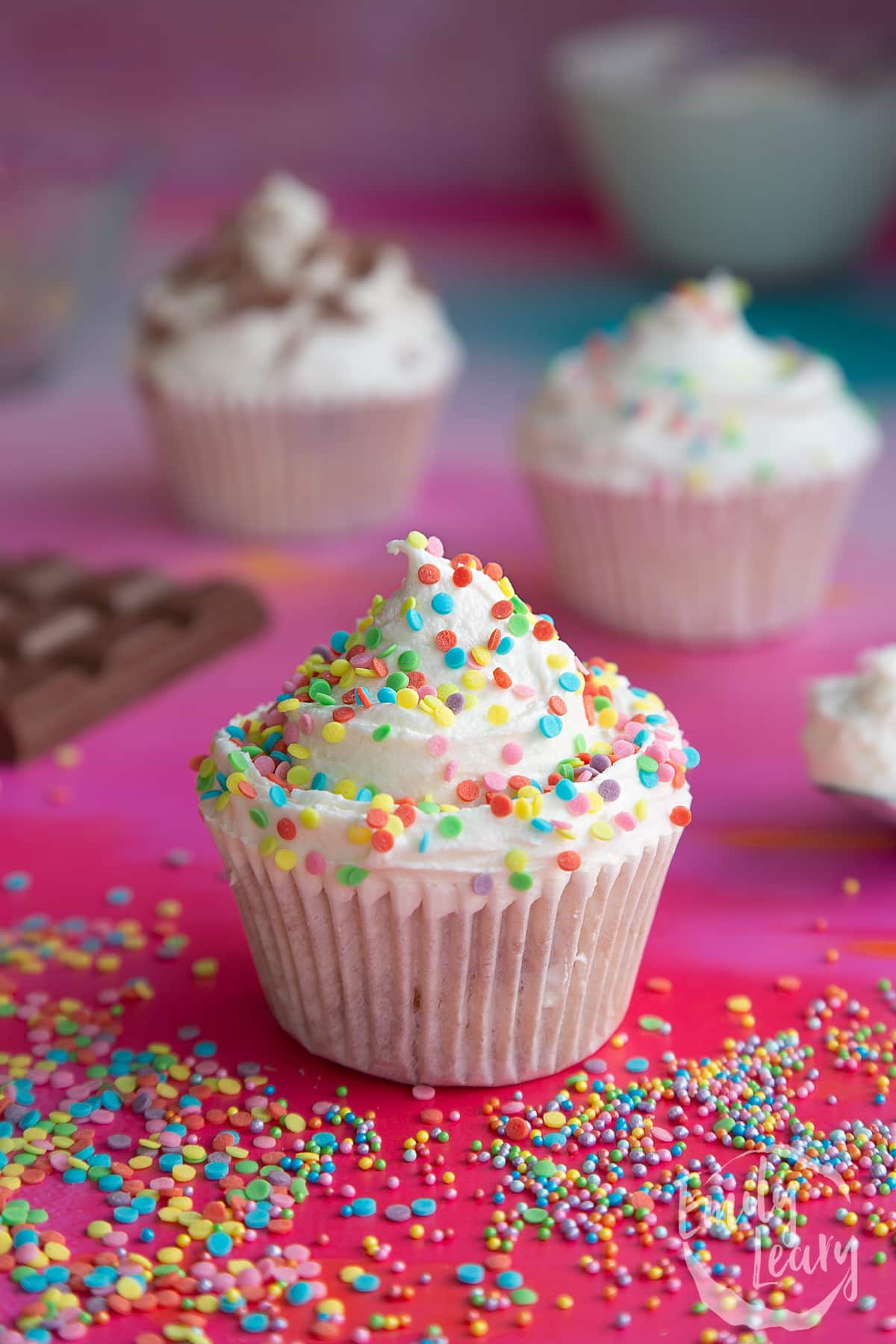 a sponge cupcake with white frosting with rainbow sprinkles on top on a red baclground surrounded by rainbow sprinkles.