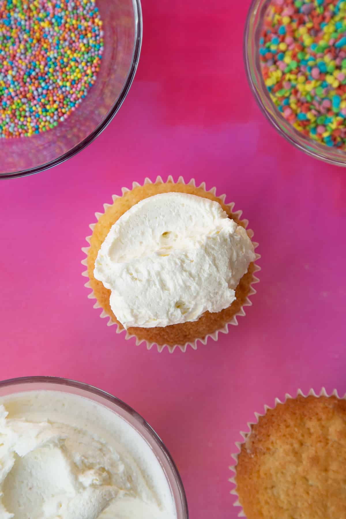 an over head view of a plain sponge cupcake white a dollop of white fosting on top on a pink background.