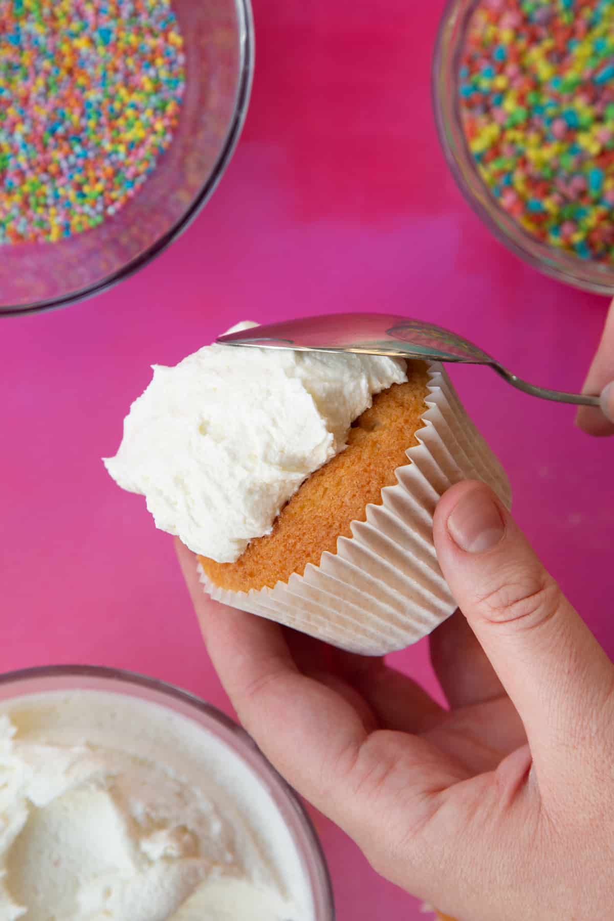 a side view of a cupcake in a hand with a spoon pushing on white frosting to the top of the cupcake.