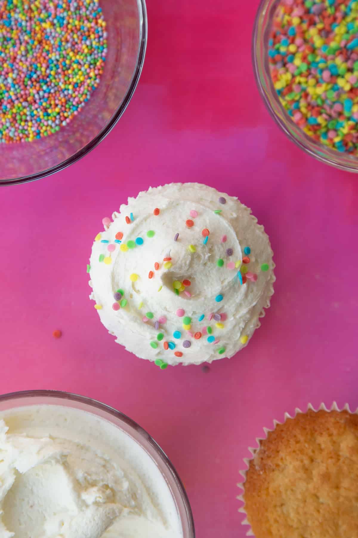 an over head view of a plain sponge cupcake with a swirl of white fosting on topped with rainbow sprinkles.