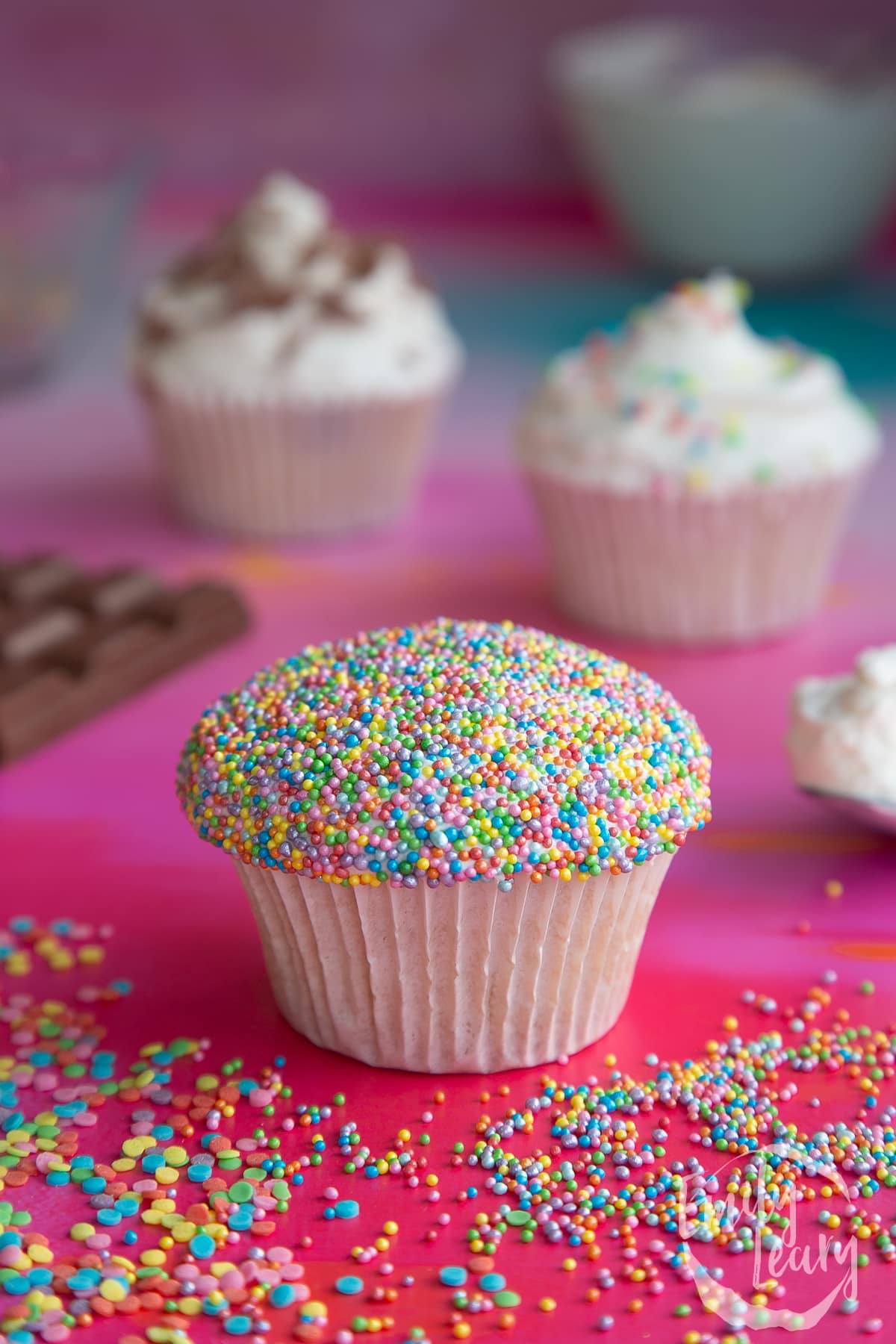 a sponge cupcake with white flat frosting with rainbow sprinkles on top on a red baclground surrounded by rainbow sprinkles.