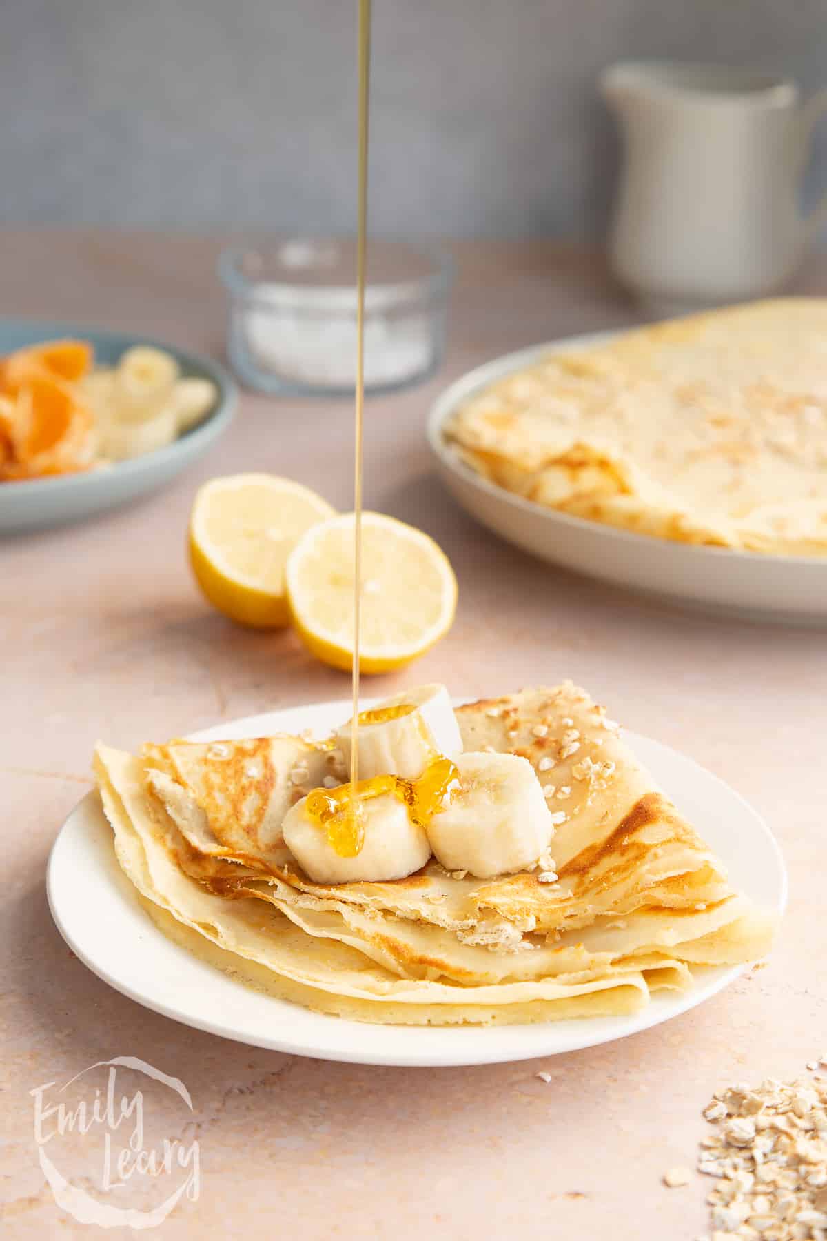 Gluten free crepe on a white plate. It is folded and topped with bananas. Golden syrup is being drizzled onto it.