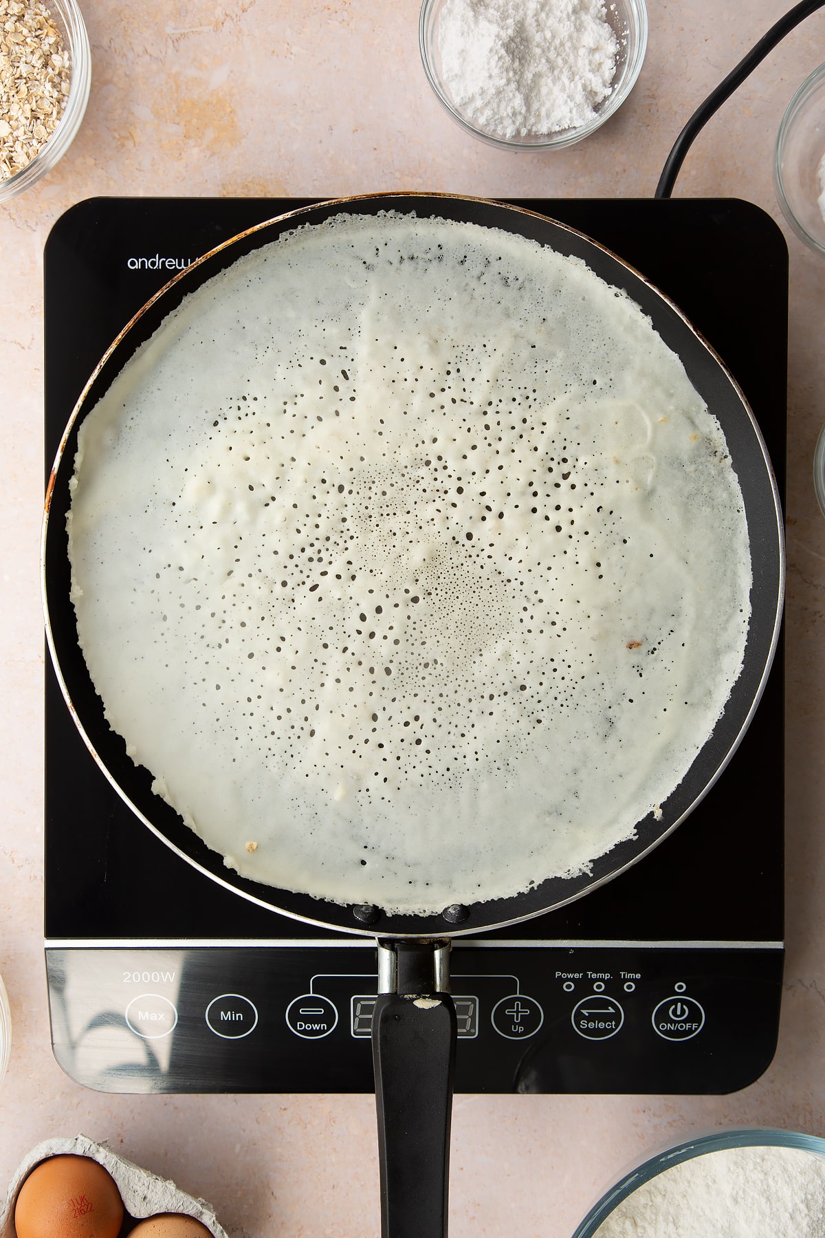 A thin layer of batter cooking in a crepe pan. Ingredients to make gluten free crepes surround the bowl.