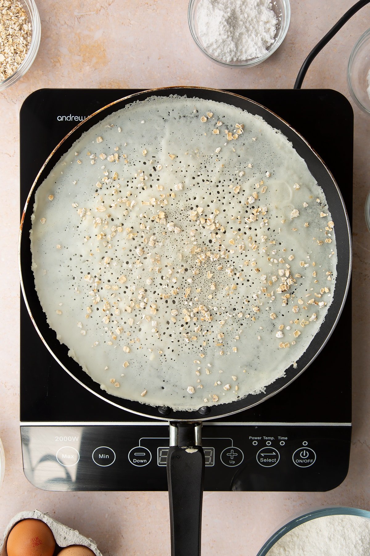 A thin layer of batter cooking in a crepe pan with oats sprinkled on top. Ingredients to make gluten free crepes surround the bowl.