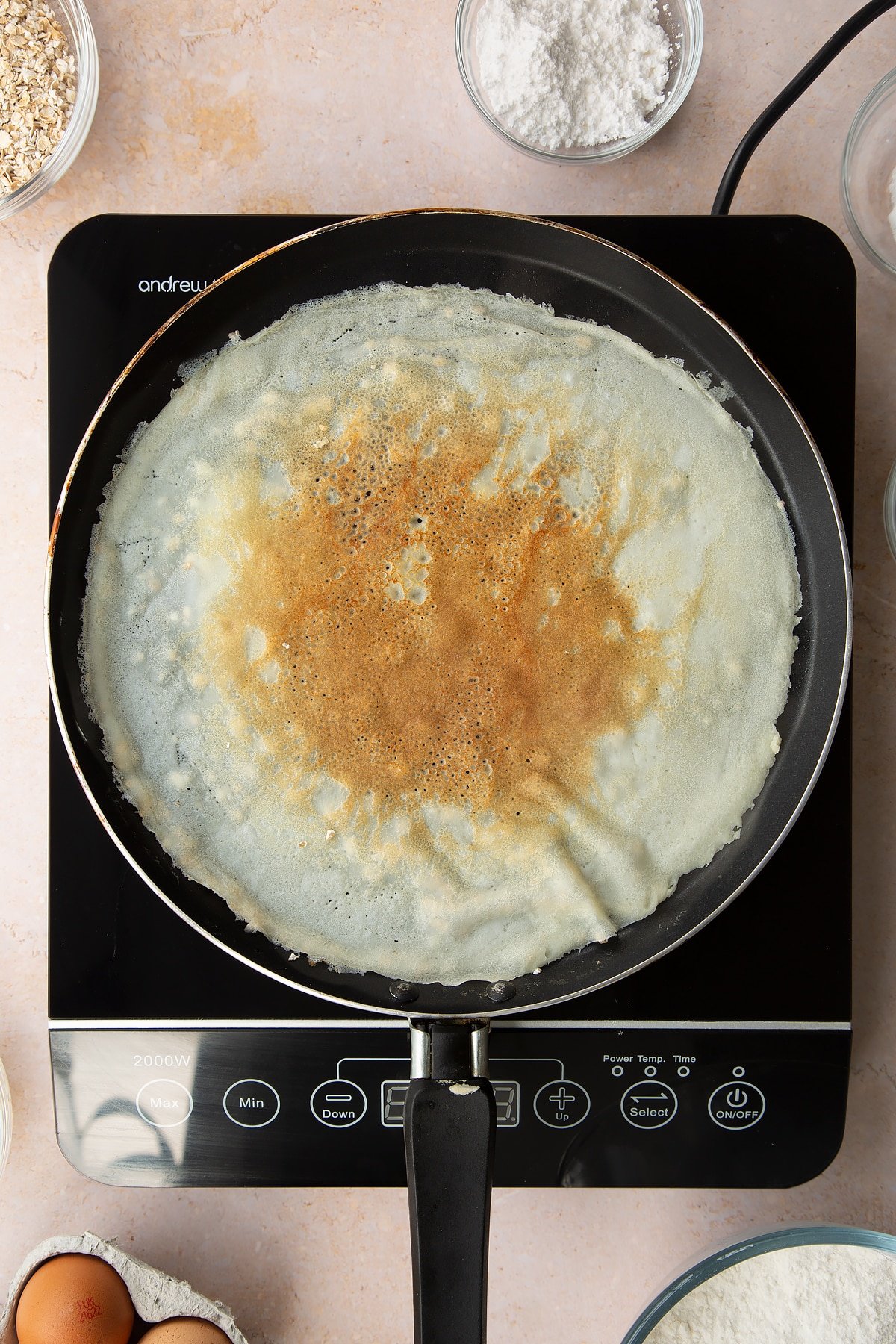 A crepe flipped over in a crepe pan. Ingredients to make gluten free crepes surround the bowl.