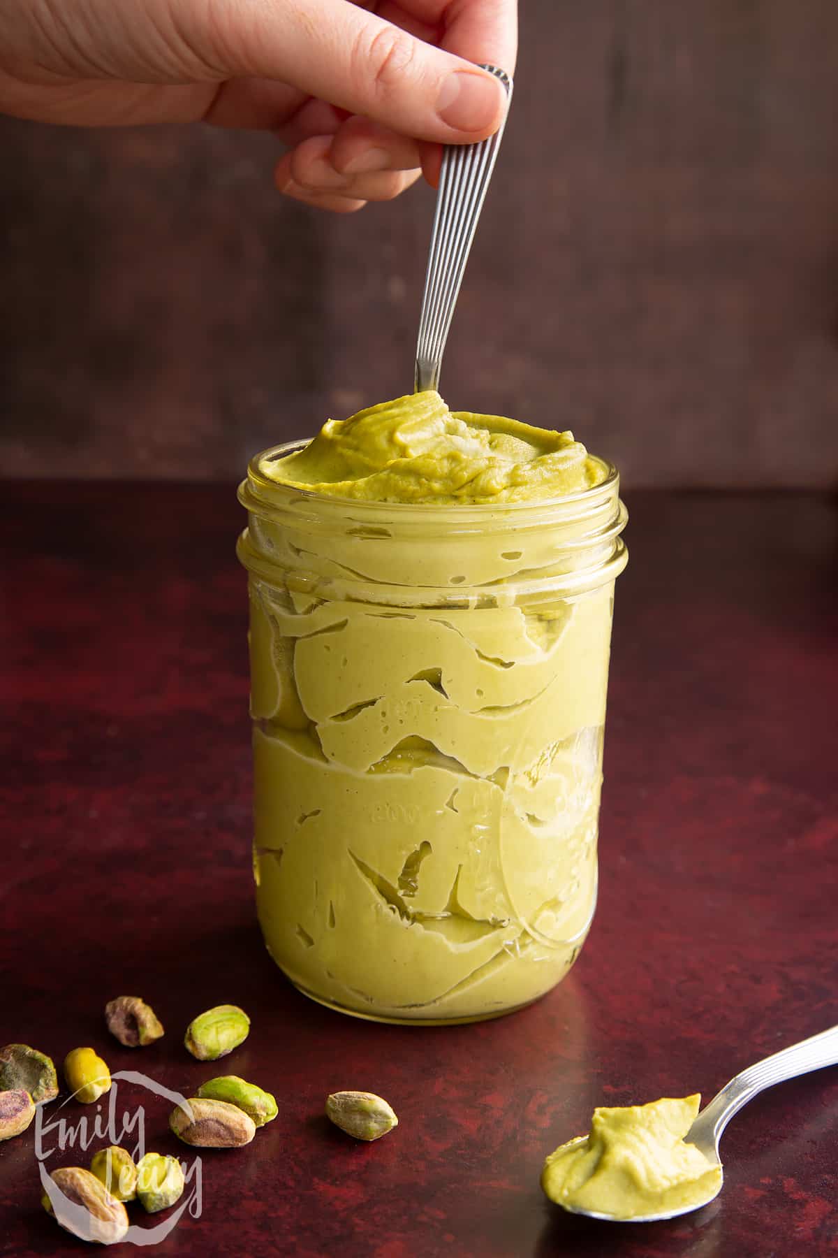 Side on shot of a jar of pistachio paste with a spoon going into the top of the jar.