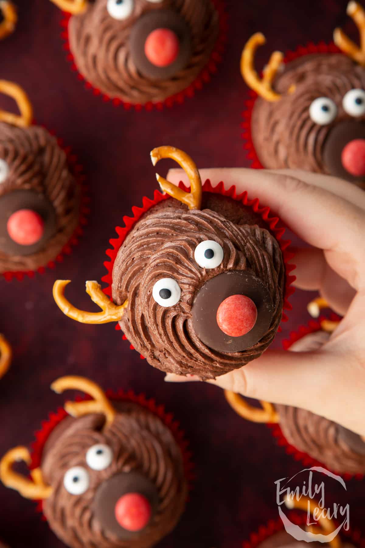 Hand holding a reindeer cupcake above more cupcakes shown from above on a dark red backdrop.