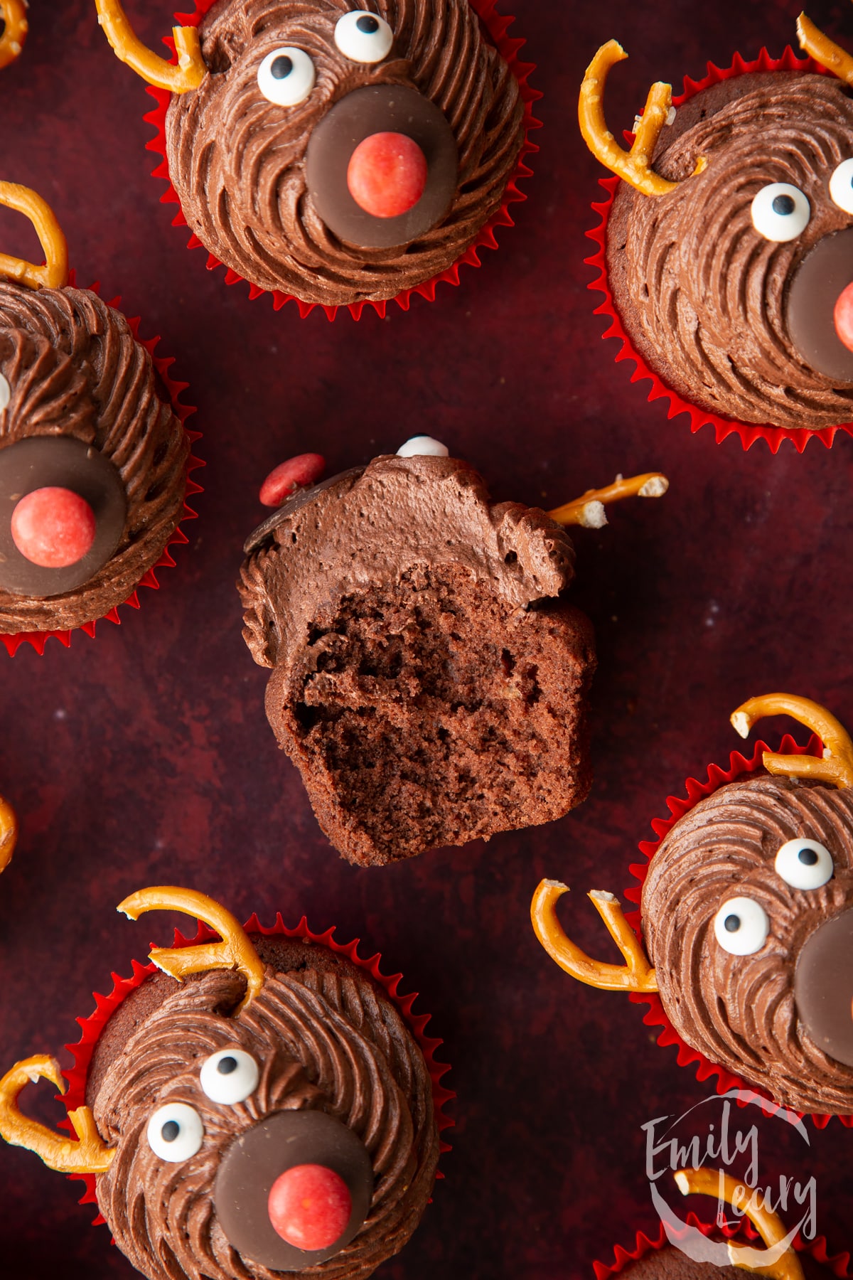 Reindeer cupcakes shown from above on a dark red backdrop. One has been bitten in half and it laying on its side.