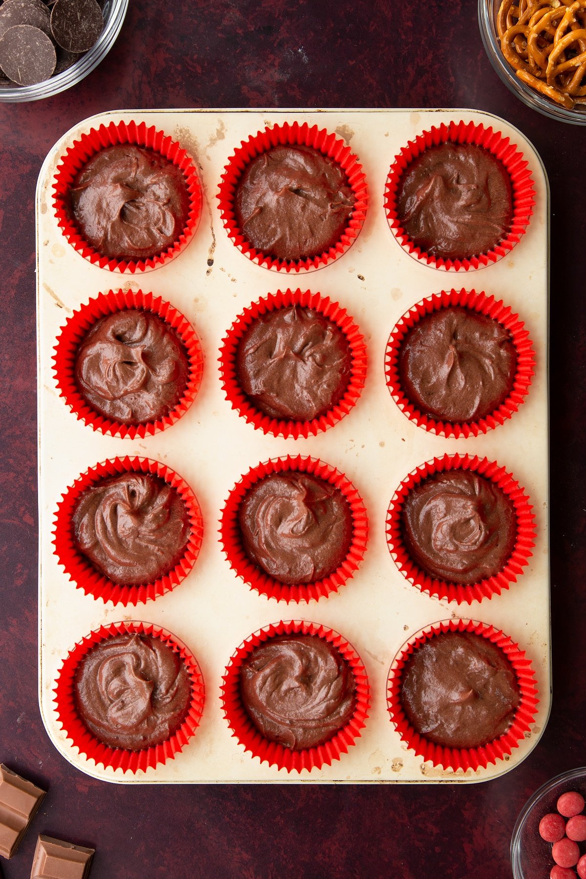 Chocolate cake batter in red cupcake cases in a tray. Ingredients to make reindeer cupcakes surround the tray.