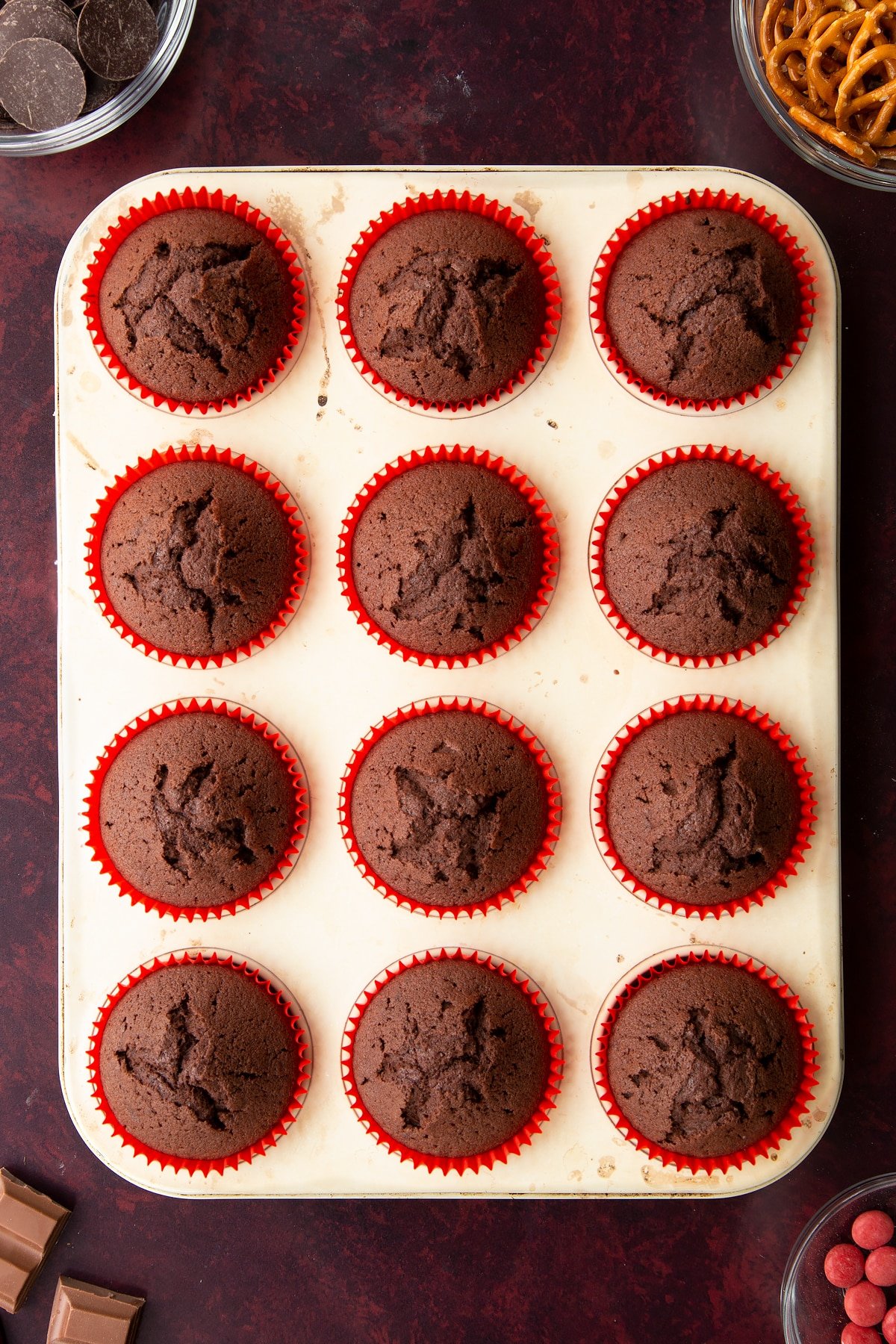Baked chocolate cakes in red cupcake cases in a tray. Ingredients to make reindeer cupcakes surround the tray.