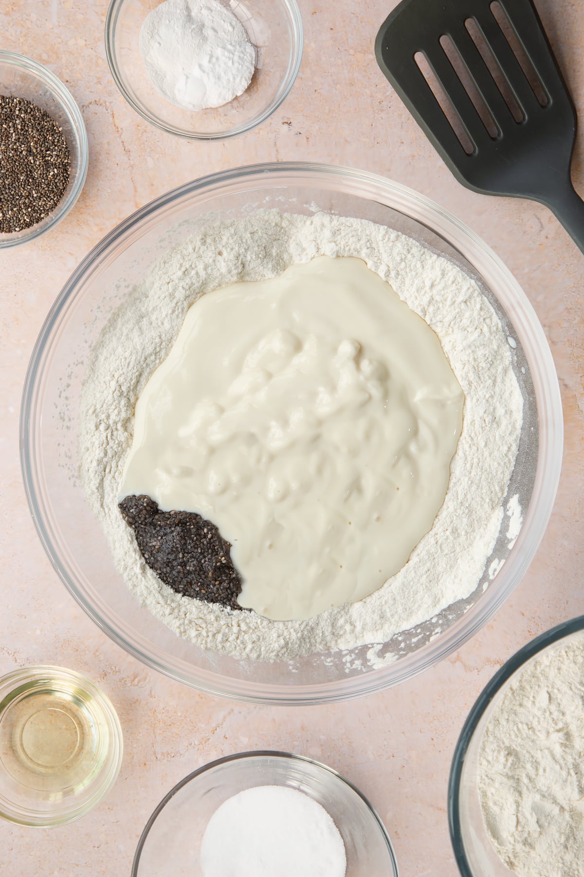 Self-raising flour, sugar, baking powder and bicarbonate of soda in a bowl. Soya yogurt, soya milk, vanilla and soaked chia seeds are in a well in the centre. Ingredients to make vegan chia pancakes surround the bowl.