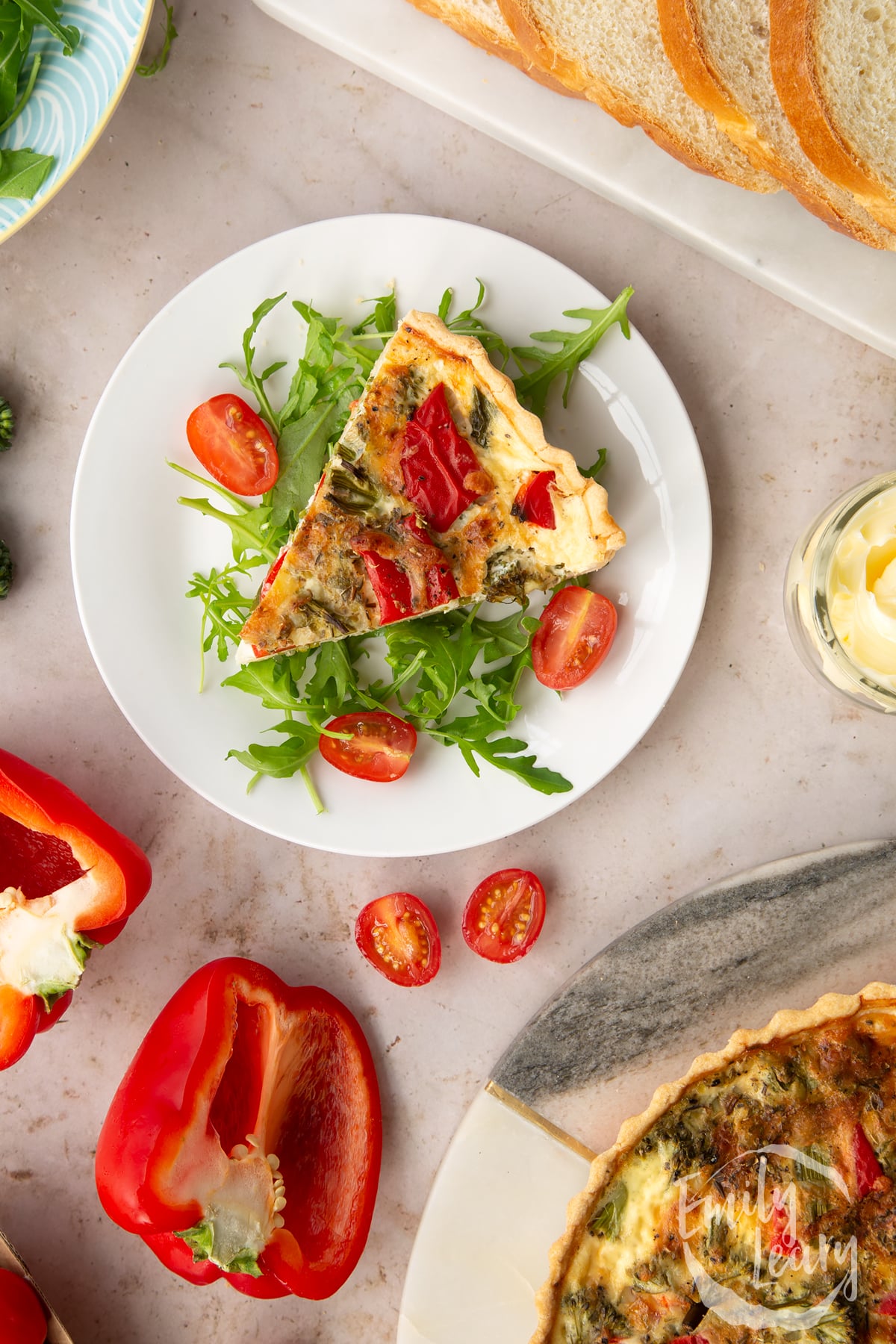 A slice of broccoli and pepper quiche served on a bed of salad on a white plate surrounded by ingredients required to complete the recipe.