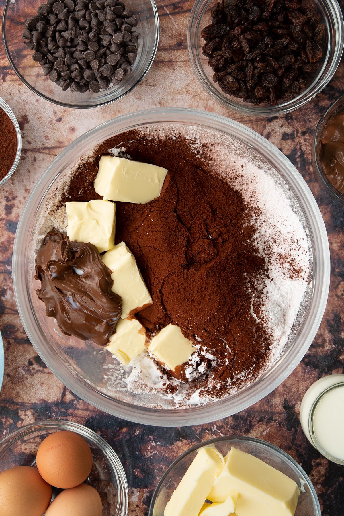 Nutella, butter, milk, cocoa and icing sugar in a bowl. Ingredients to make chocolate raisin muffins surround the bowl.
