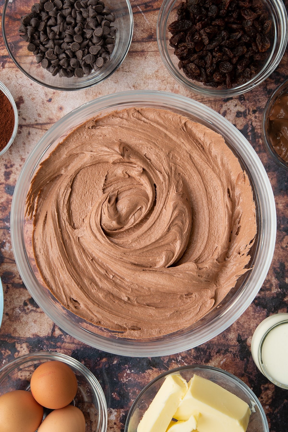 Nutella buttercream in a bowl. Ingredients to make chocolate raisin muffins surround the bowl.