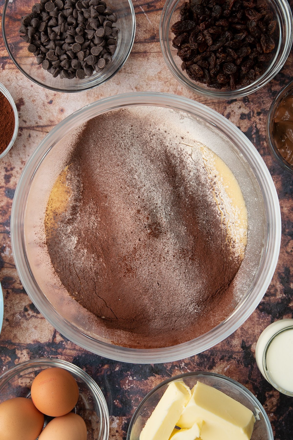 Butter, oil, sugar and eggs in a bowl with flour and cocoa on top. Ingredients to make chocolate raisin muffins surround the bowl.