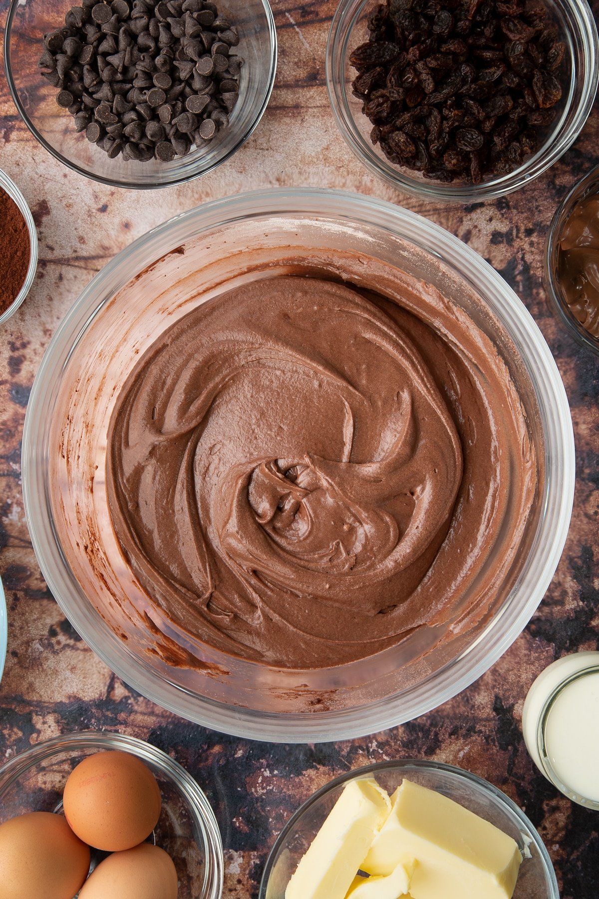 Chocolate cake batter in a bowl. Ingredients to make chocolate raisin muffins surround the bowl.