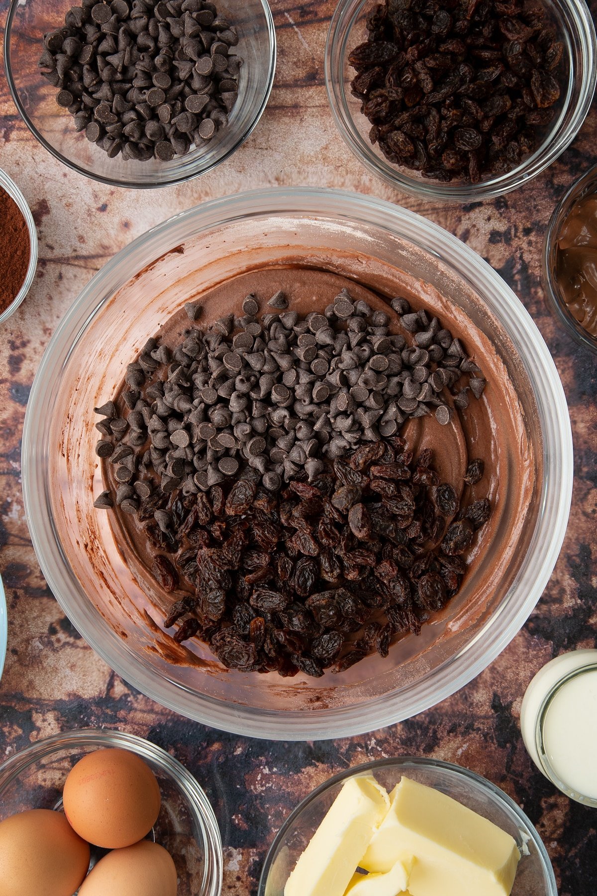 Chocolate cake batter in a bowl with raisins and dark chocolate chips on top. Ingredients to make chocolate raisin muffins surround the bowl.