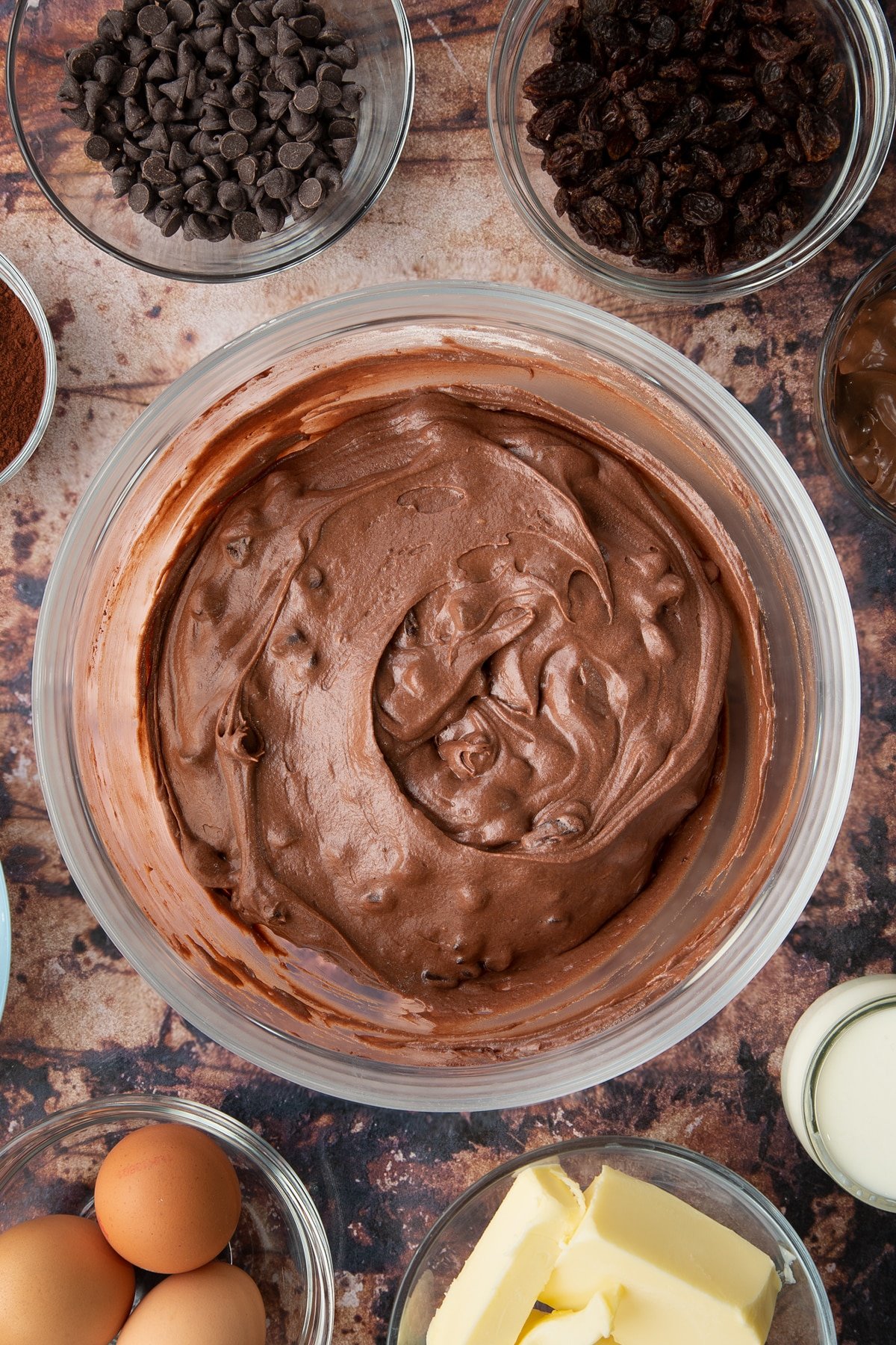 Chocolate raisin muffin batter in a bowl. Ingredients to make chocolate raisin muffins surround the bowl.