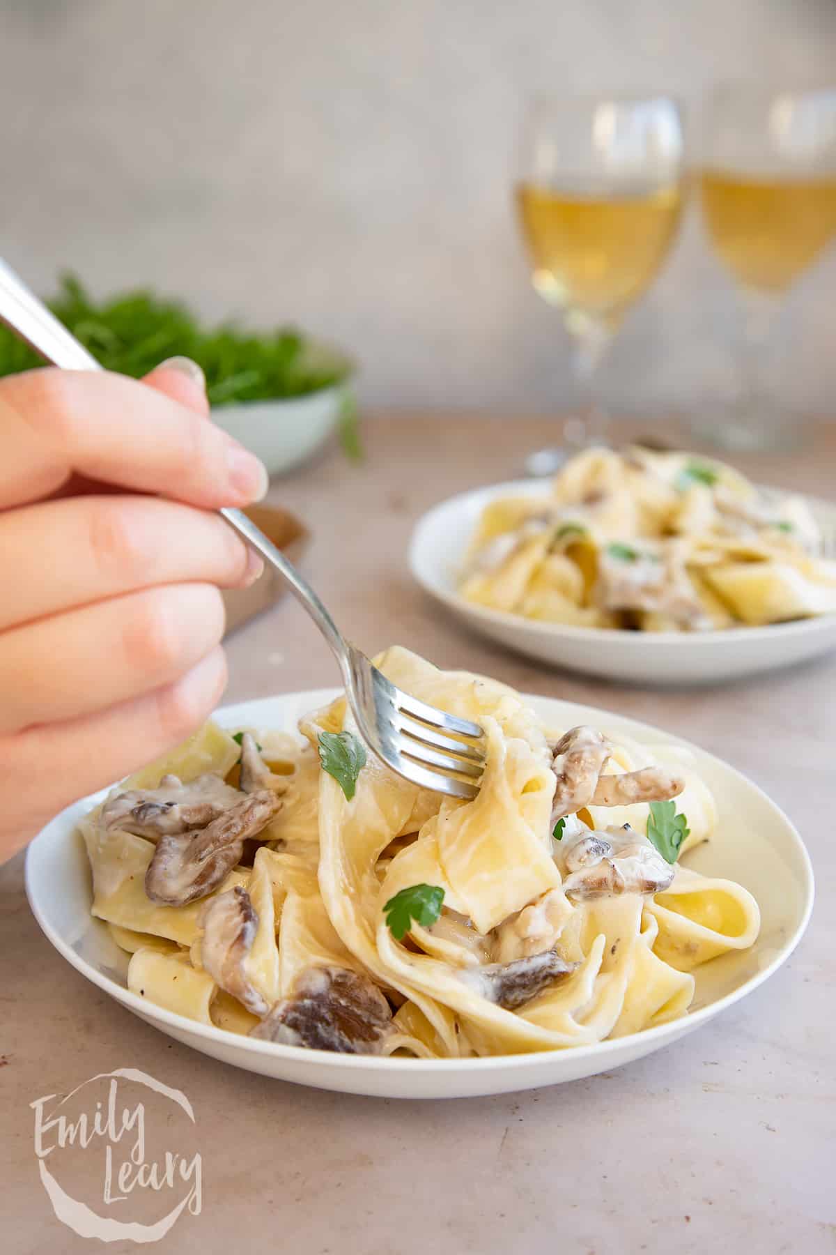 side view of Creamy mushroom pappardelle topped with parsley in a white pasta bowl with a hand holding a fork with a string of pasta.