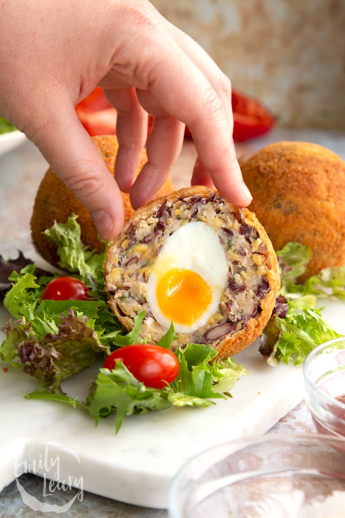 side view of a hand holding a cut vegetarian scotch egg on a bed of salad.