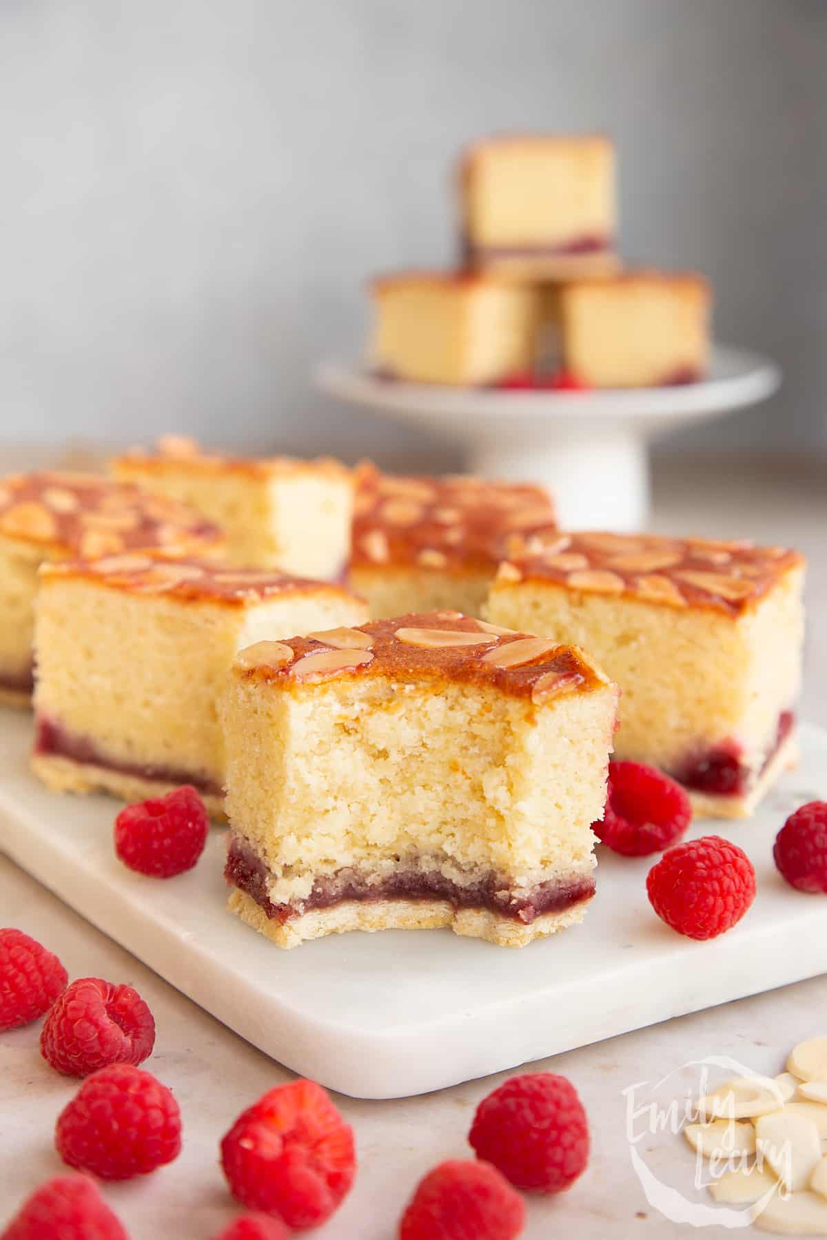 Squares of Bakewell tray bake on a white marble board. One has a bite out of it.
